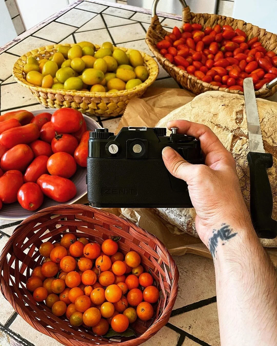 A person holding a camera at a table with baskets and plates of ripe tomatoes of various colors, including red, yellow, and orange, with a loaf of bread and a knife also on the table.