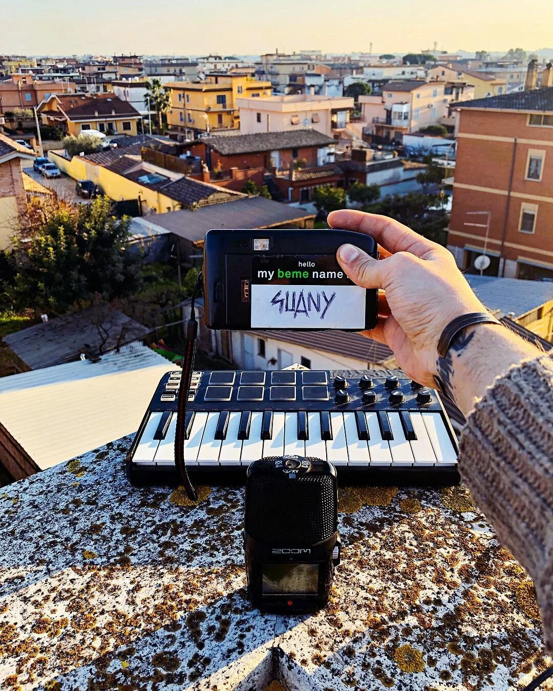 A person holds a smartphone with handwritten text 'SUANY' on the screen, background of a cityscape with multiple rooftops, a keyboard, and a voice recorder on a rooftop ledge during sunset.