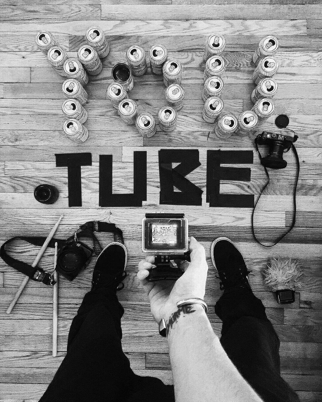 The photo shows a person's legs and feet, with various photography gear, including cameras and a drone, on a wooden floor. In front of the person, there are stacked cans arranged to spell 'TRU'. The cans have pull tabs on top. Below the cans, large b