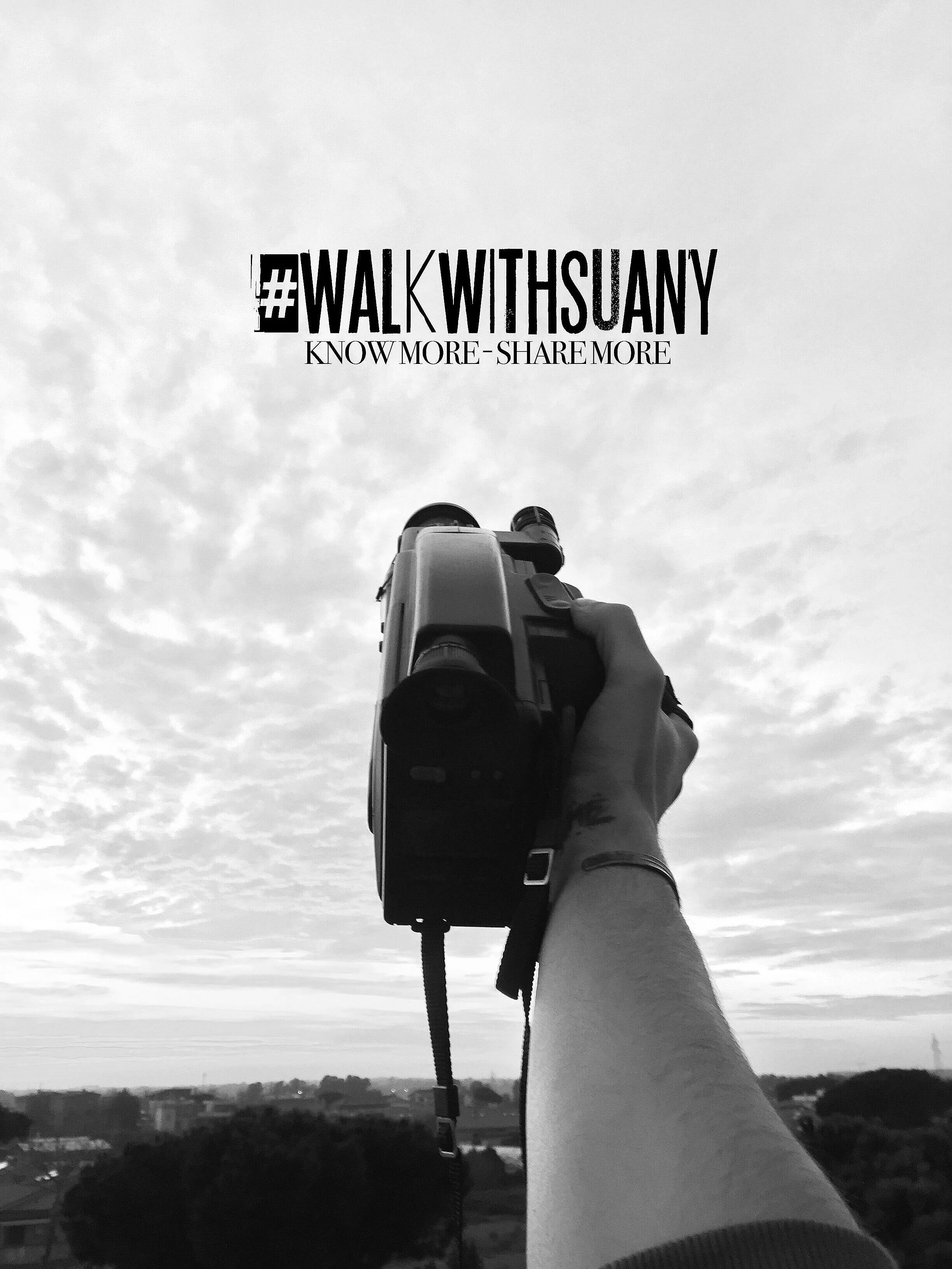 A person holding a camera pointed upward toward the sky, with a hashtag #WALKWITHSUANY and the text "KNOW MORE - SHARE MORE" in the sky.