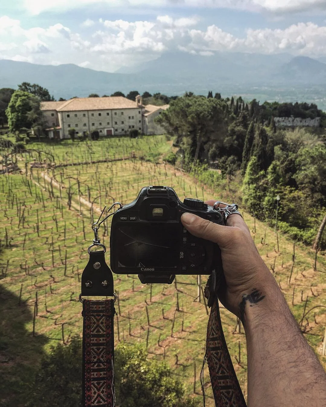 A person is holding a Canon camera, taking a photo of a vineyard with a large building in the background and mountains under a partly cloudy sky.