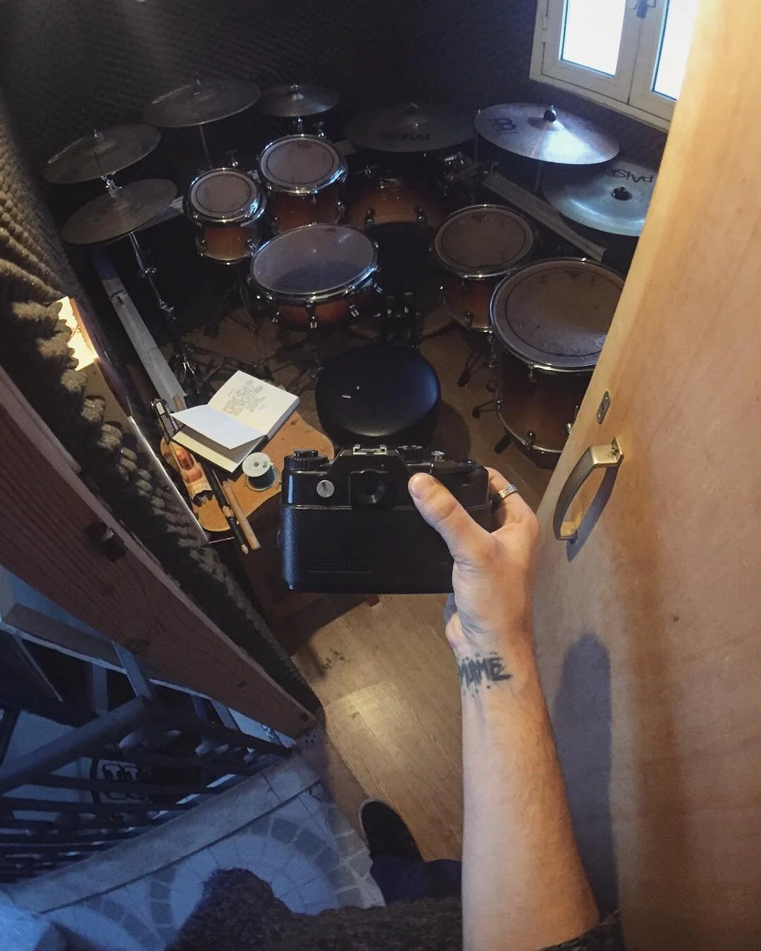 A person taking a selfie in a mirror showing a drum set, a notebook, and a musical note on a paper in a small music room.