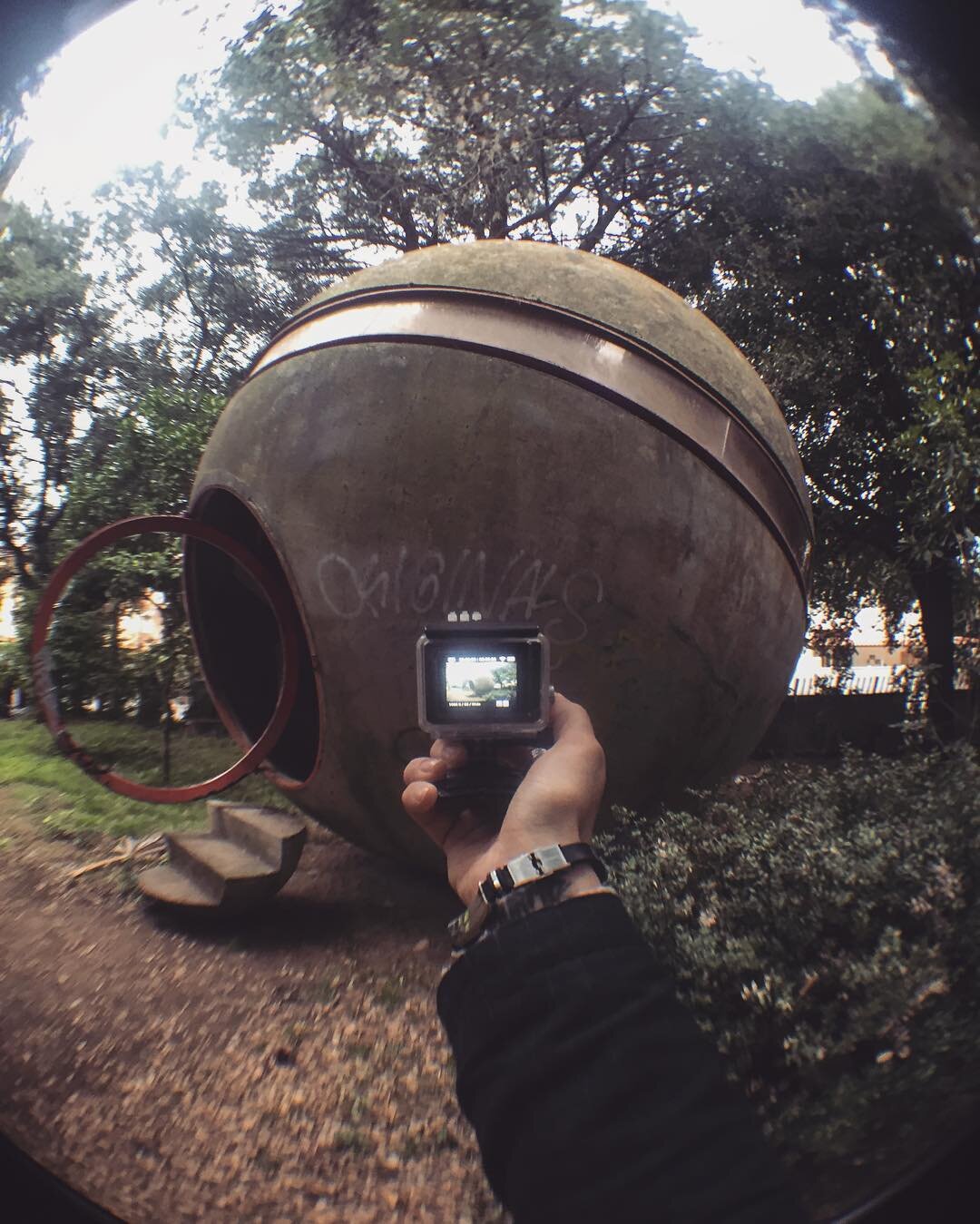 Person taking a fisheye photo of a large, upside-down, old bowling ball sculpture outdoors, surrounded by trees and grass.
