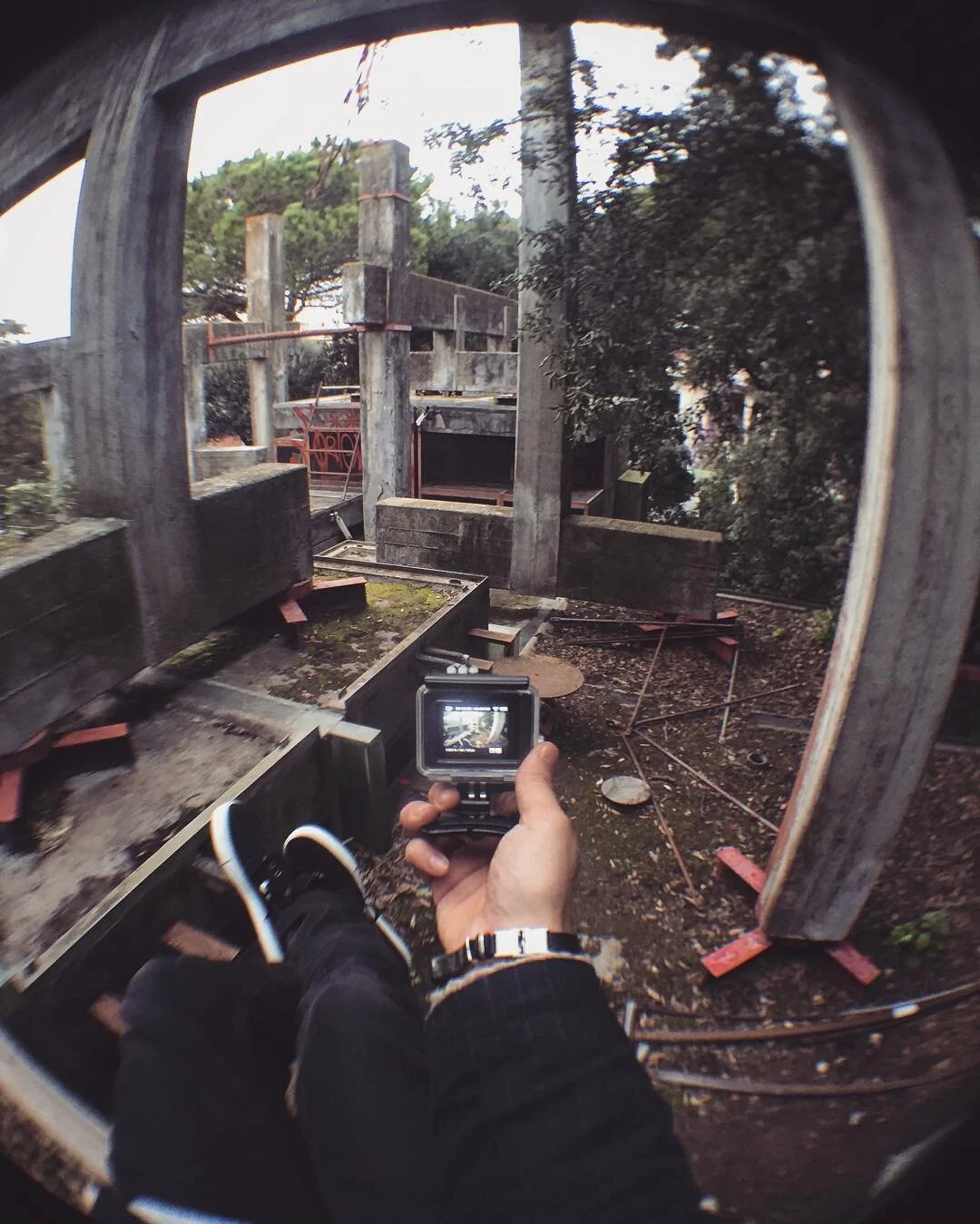 Person holding a camera, taking a photograph of an abandoned, weathered structure with concrete and metal elements, surrounded by trees.