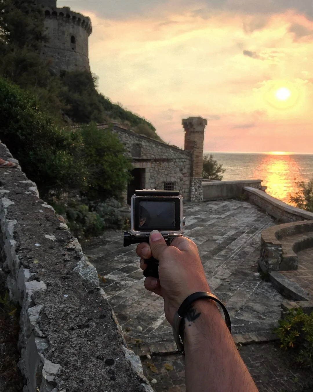 Person taking a photo with a camera of a sunset over the ocean, with stone castle towers and walls in the background.