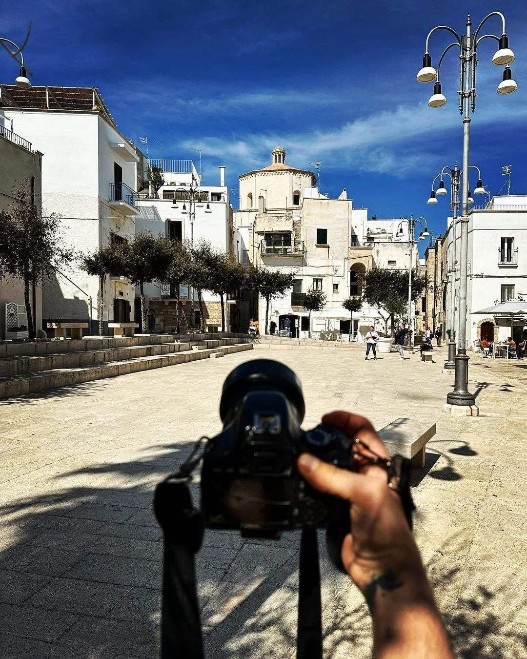 A person holding a camera taking a photo of a historic Mediterranean town square with white buildings, trees, and street lamps on a sunny day.