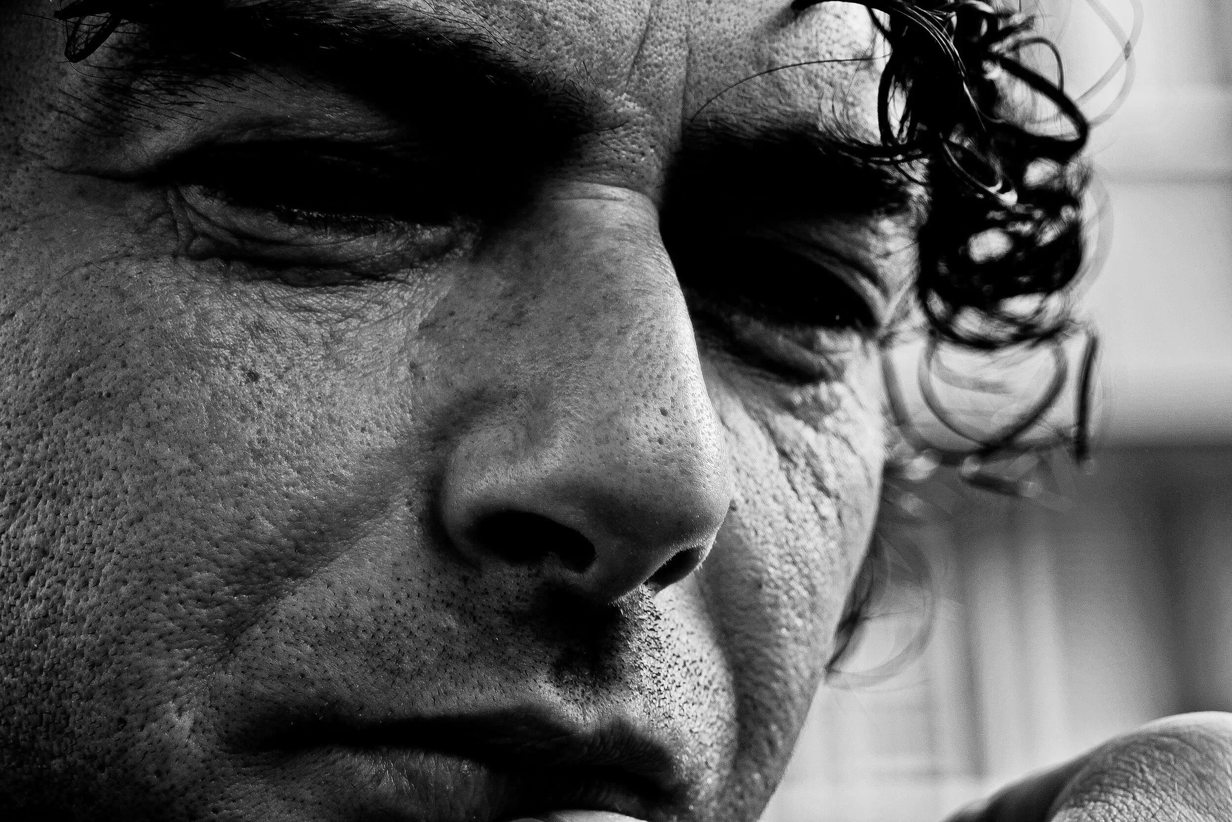 Close-up black-and-white photograph of a person's face, highlighting textured skin, closed eye, and curly hair.