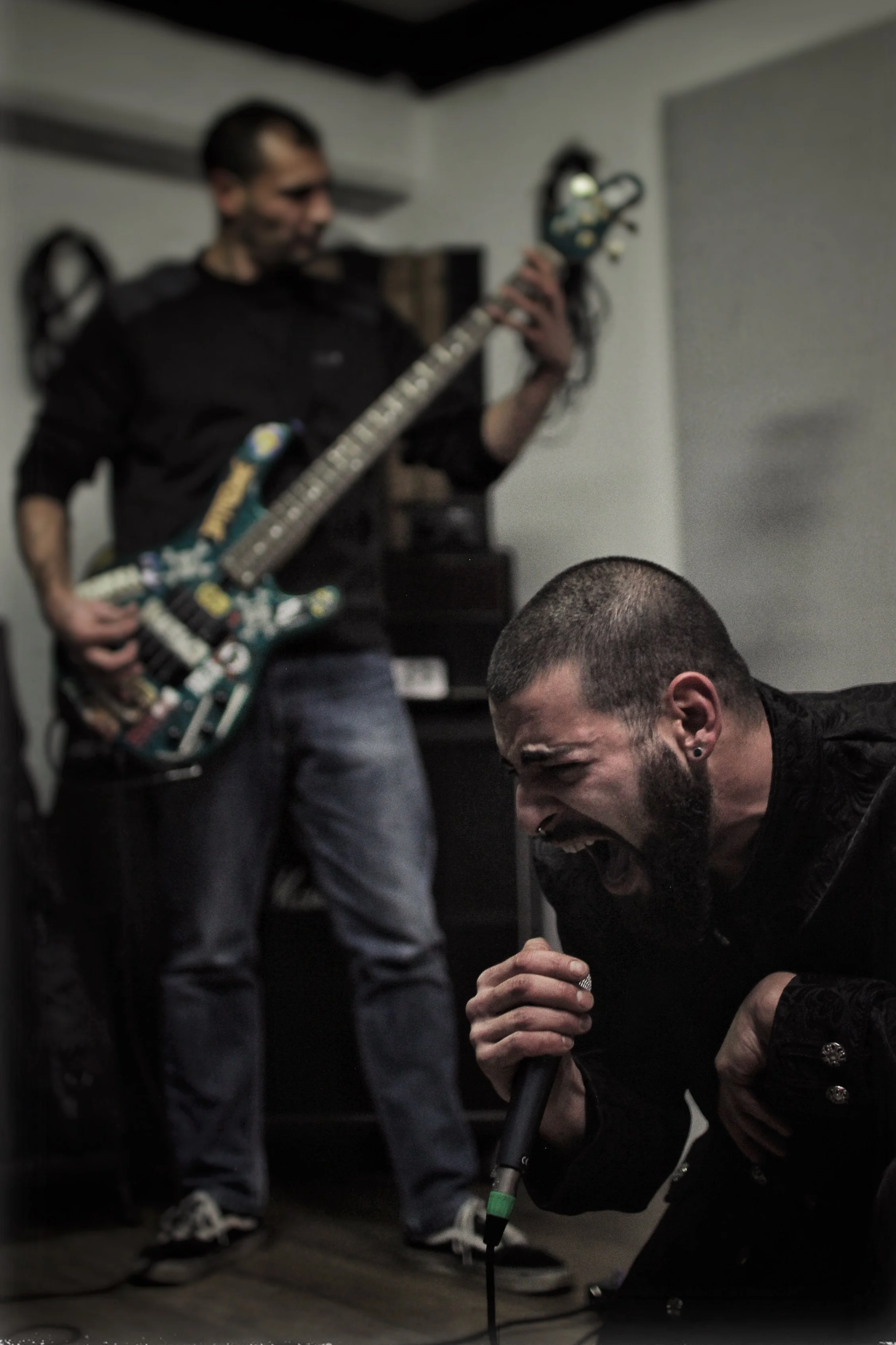 A man with a beard and earrings kneels on the floor holding a microphone, while another man with short hair and a tattoo on his neck plays a guitar in a room with white walls.