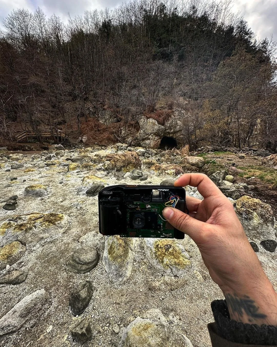 Person holding a camera outdoors near a rocky stream with a cave entrance in the background, surrounded by leafless trees and hills.