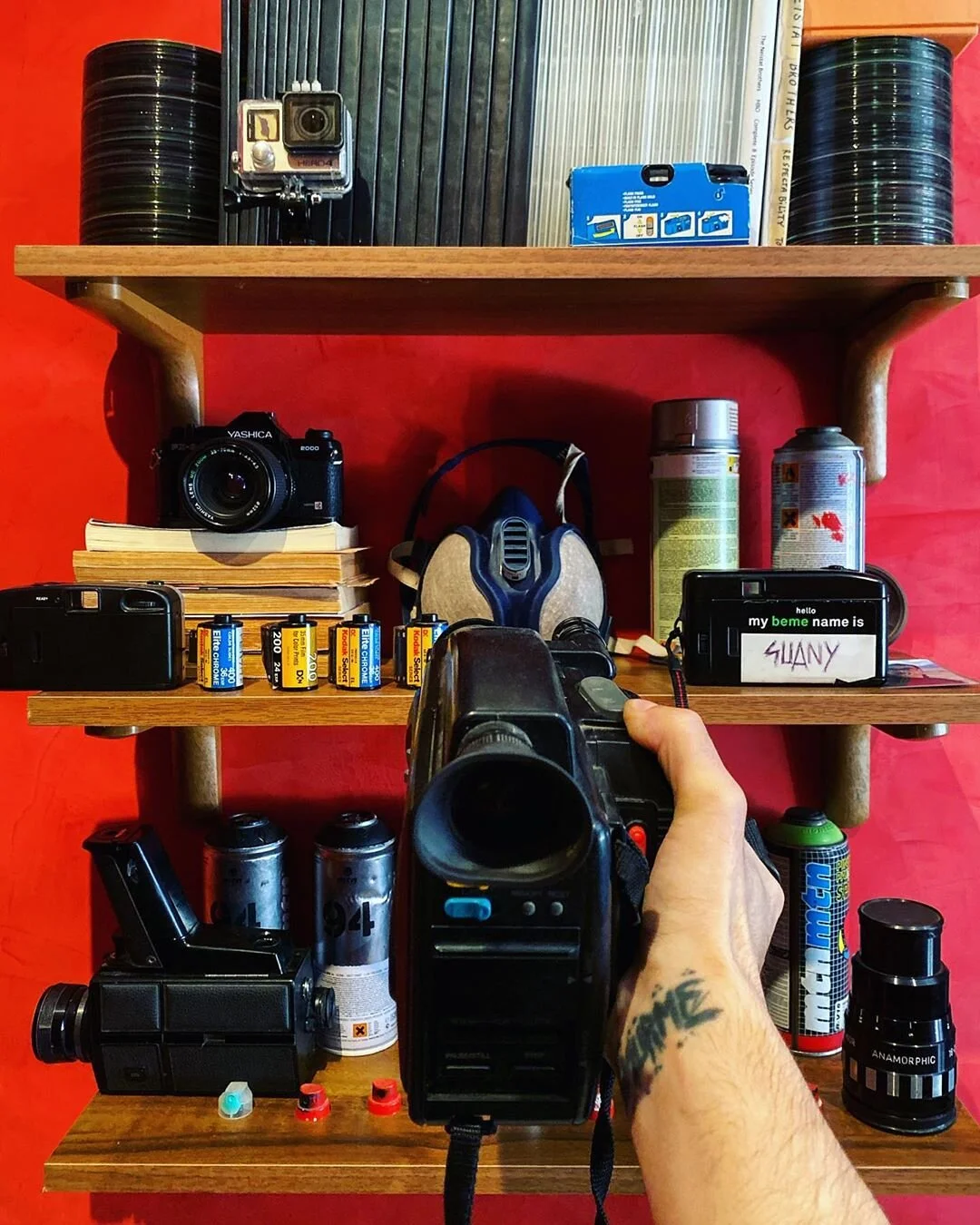 A wooden shelf with various camera equipment, film canisters, and a person's hand holding a camera in the foreground.