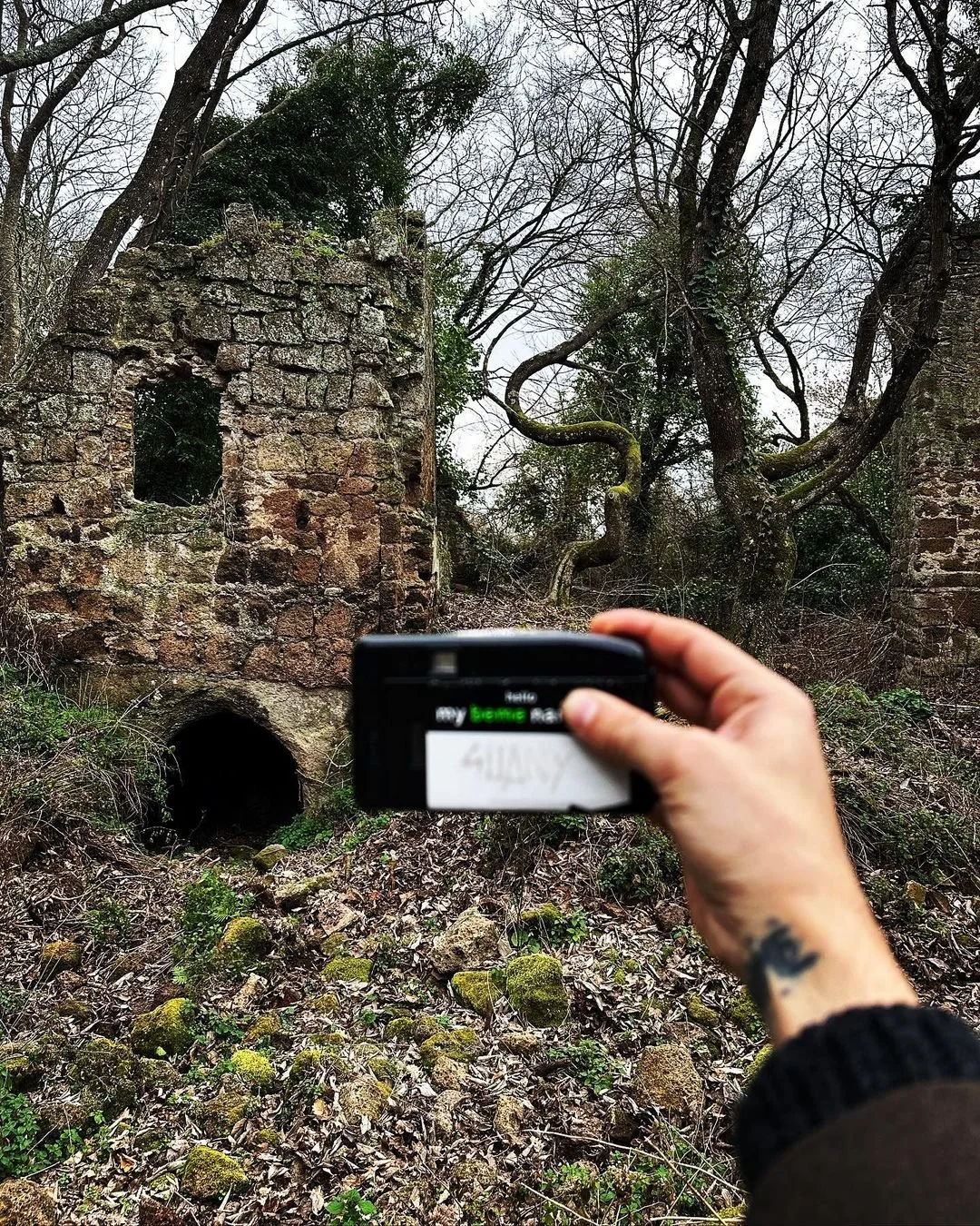 A person holding a camera in front of an old, abandoned stone structure with arched entrance, surrounded by leafless trees and overgrown vegetation.