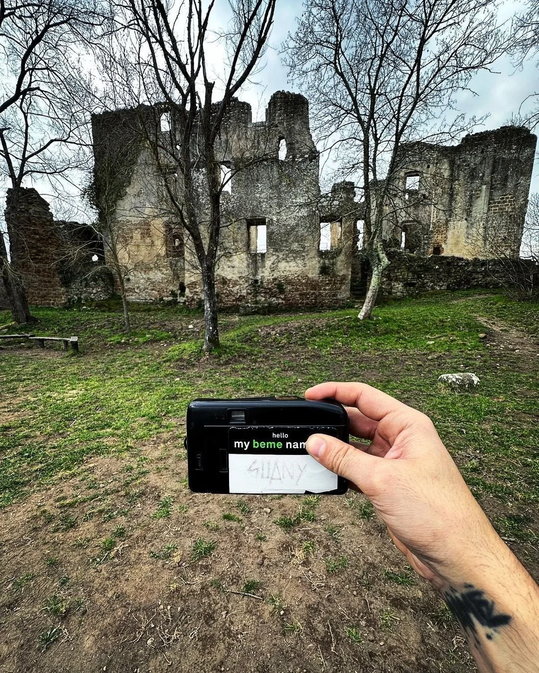 Person holding a camera with a note that says 'SHANY' in front of a weathered stone castle surrounded by leafless trees and a partly grassy, dirt ground.