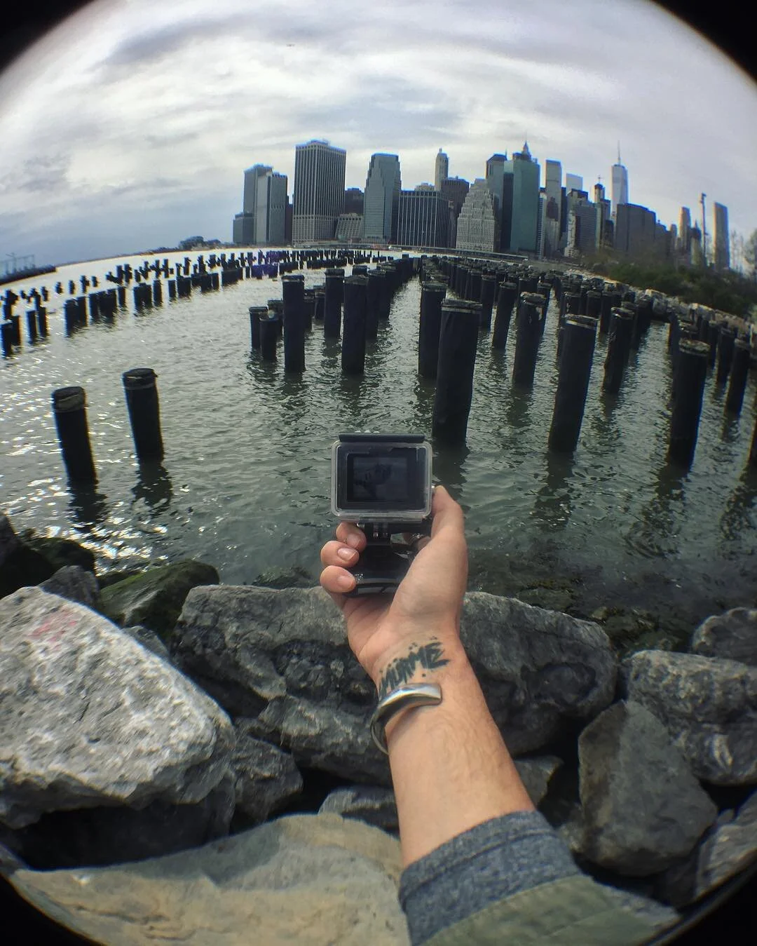 A person holding a GoPro camera takes a photo of the New York City skyline and water pier posts, with the city in the background and a cloudy sky overhead.