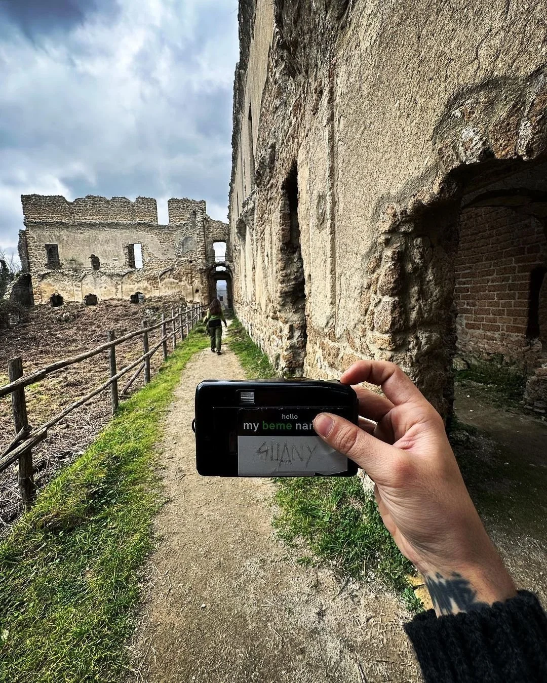 A person taking a photo of an ancient stone castle ruins with a smartphone, along a dirt path bordered by a wooden fence, under cloudy skies.