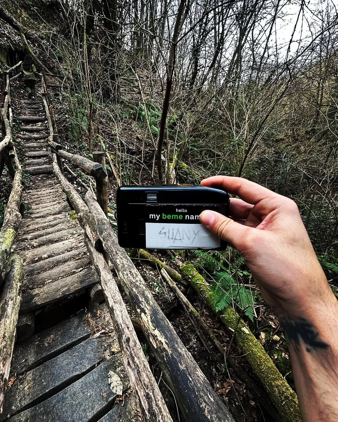 A person holds a camera with a paper note reading 'SHANY' in front of a forest trail with a wooden bridge and a person walking up the stairs in the background.