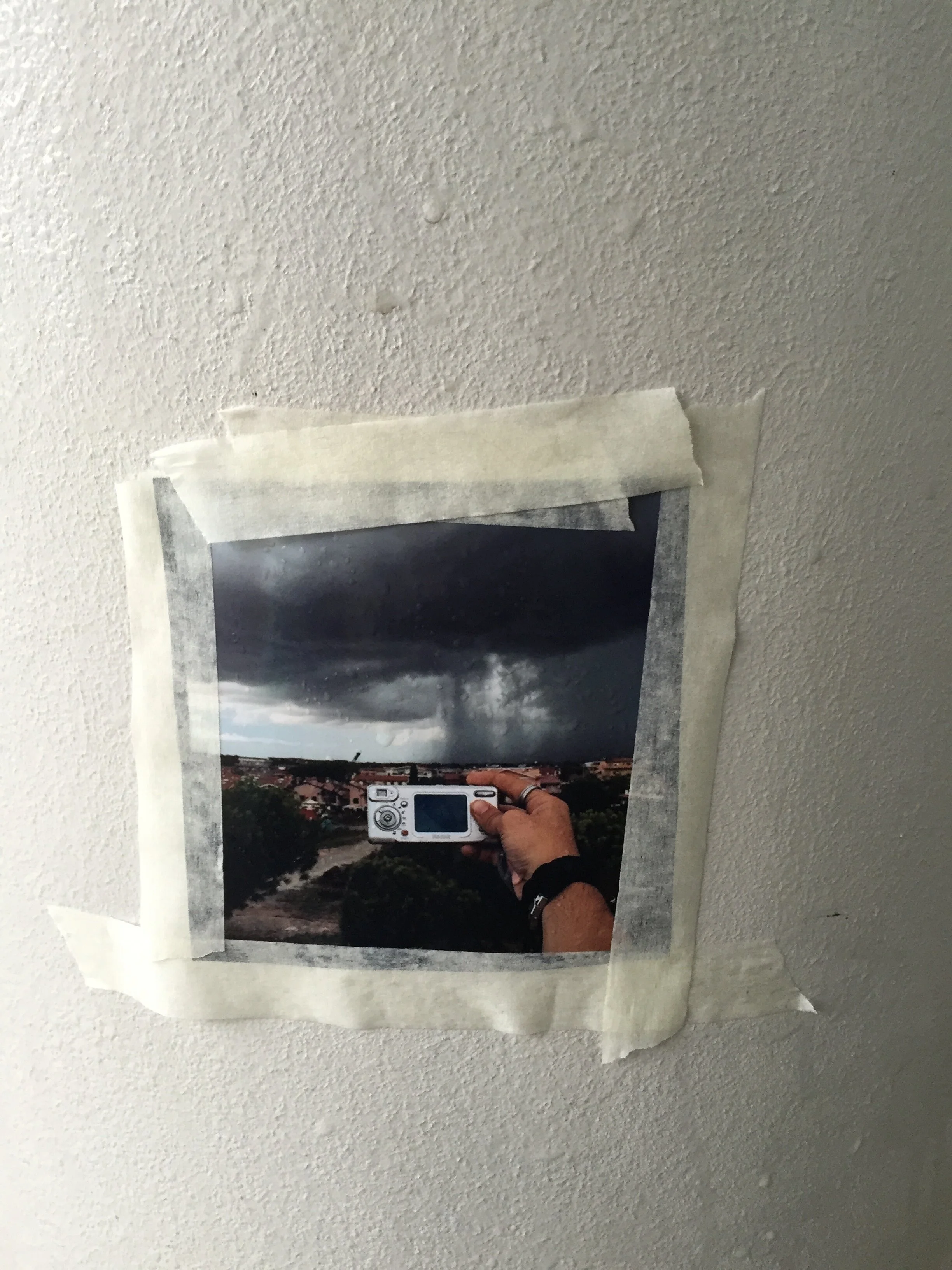 A photograph of a printed photo taped to a wall, capturing a person holding a camera and taking a picture of a landscape scene with houses, trees, and a cloudy sky.