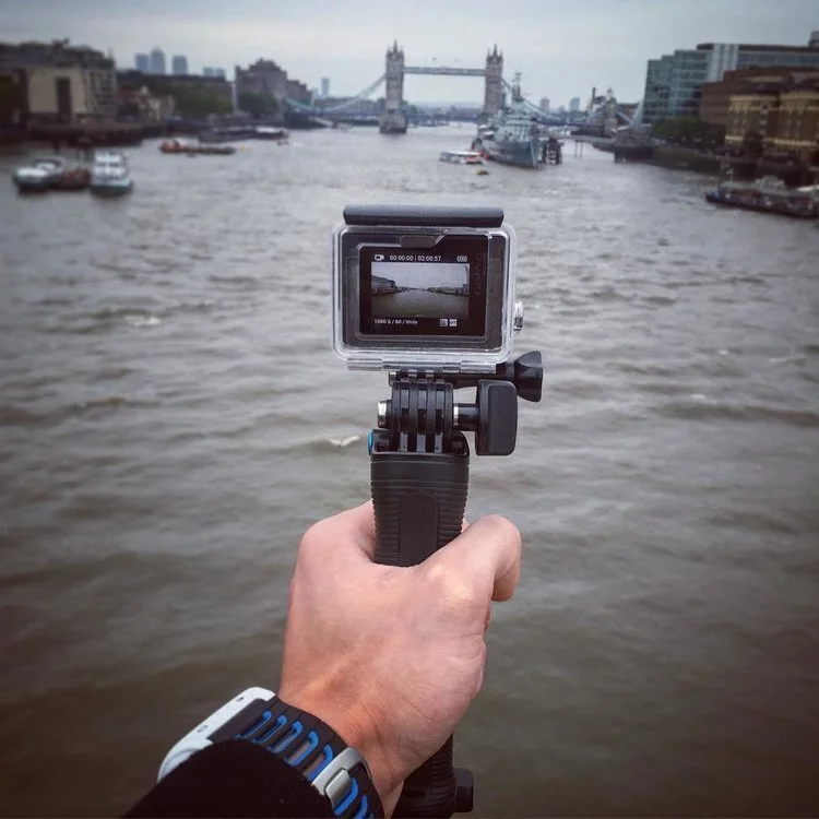 A person holding a camera on a selfie stick, capturing the Tower Bridge and River Thames in London during daytime.