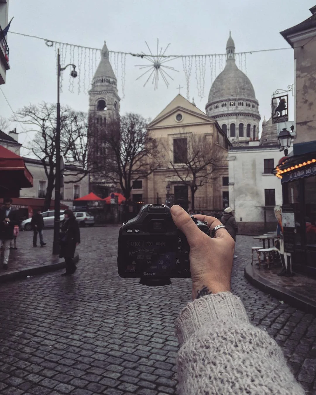 A person holding a Canon camera takes a photo of the Sacré-Cœur Basilica in Paris, with cobblestone street and trees in the foreground, and cloudy sky overhead.
