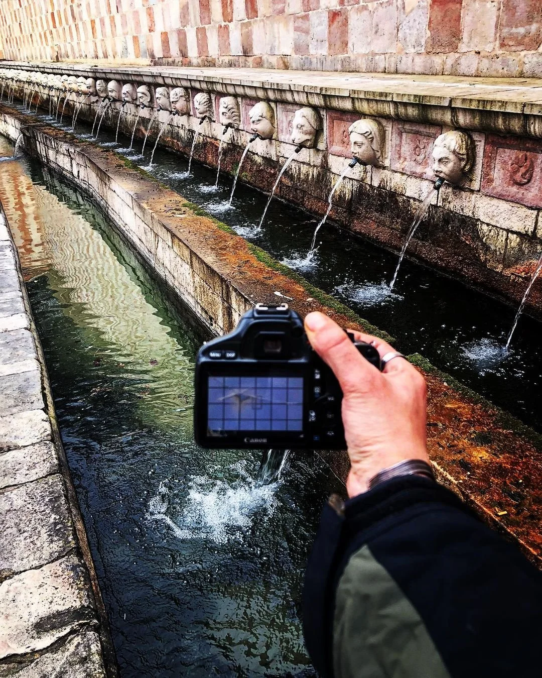 Close-up of a person holding a camera near a stone fountain with water streams coming out of carved heads on a brick wall.