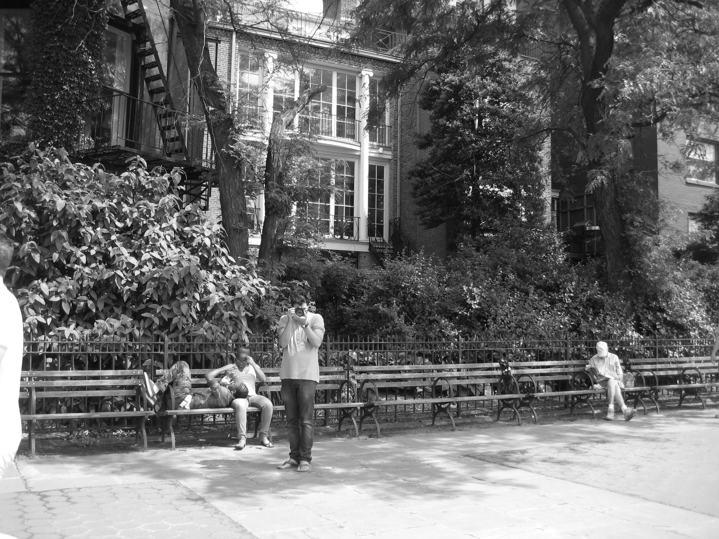 Black and white photo of a park scene with a row of benches along the sidewalk. Several people are sitting on the benches, and one person is standing with a camera. There are trees with dense foliage behind the benches and a large brick building with