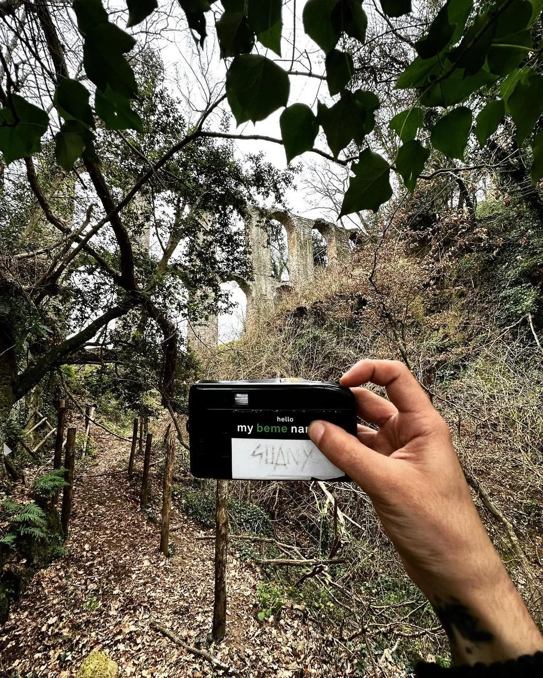 A person holds a small black device with a handwritten note reading 'SHANNON' in a forested area. Behind, a trail with a wooden fence runs through dense trees and shrubs, and large stone arches are visible in the background.