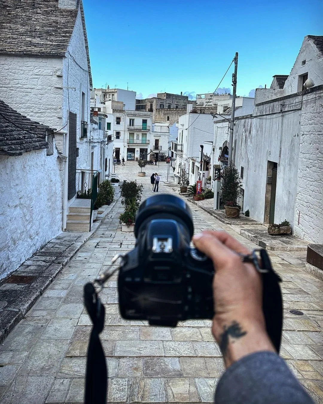 A person holding a camera takes a photo of a small white town square with buildings on both sides, some with potted plants, and a few people standing and walking in the distance. The sky is clear and blue.