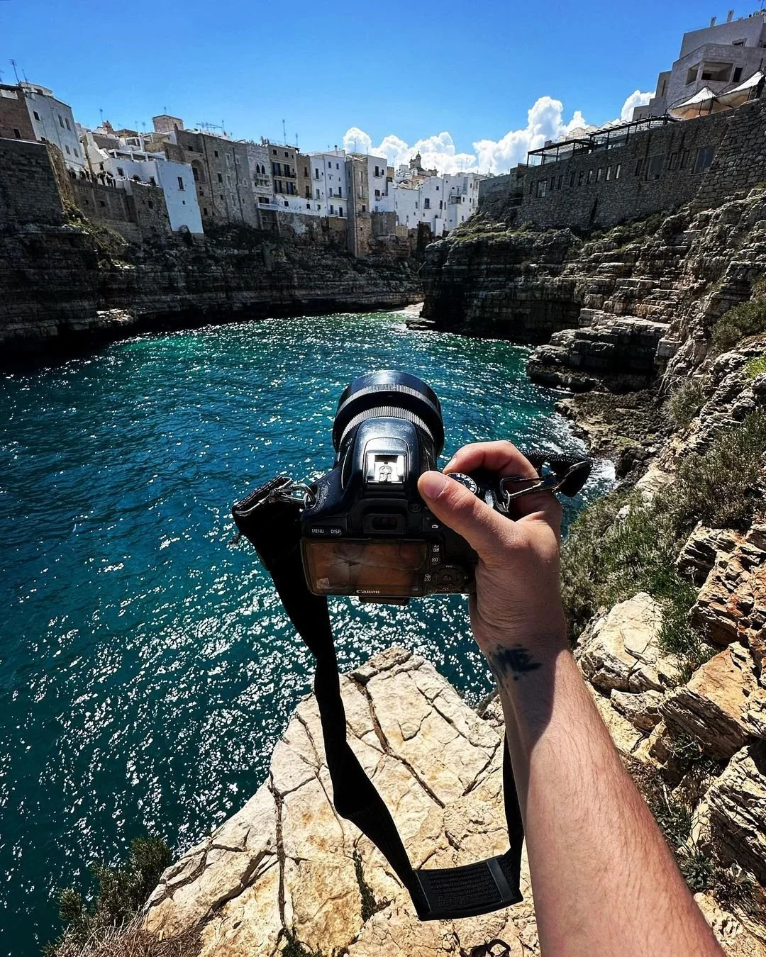 A person holding a camera while standing on rocky cliffs overlooking a blue water cove with white buildings on cliffs in the background.