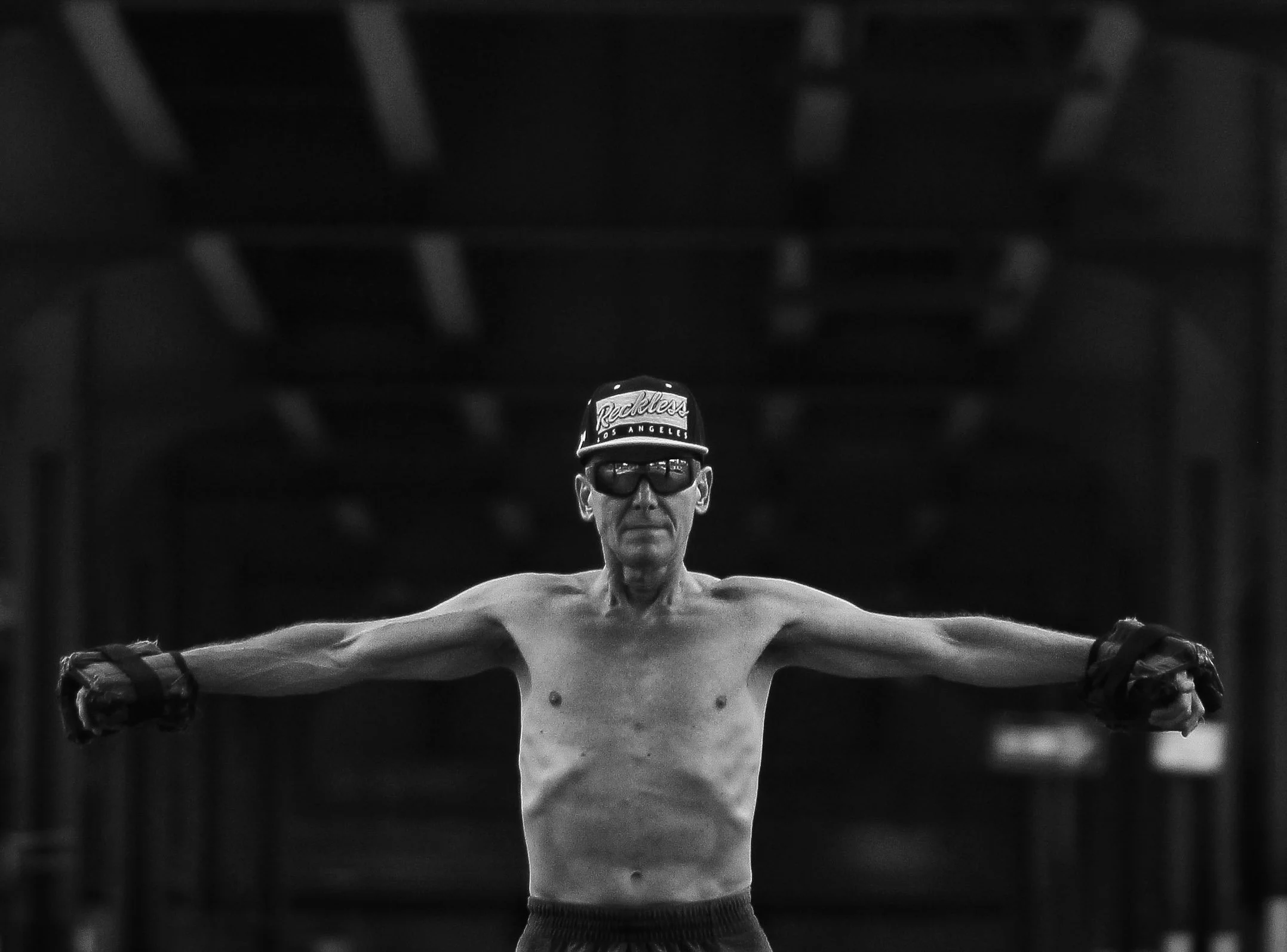 Black and white photo of a shirtless male boxer with cap and sunglasses, standing with arms extended, in a training or competition setting.