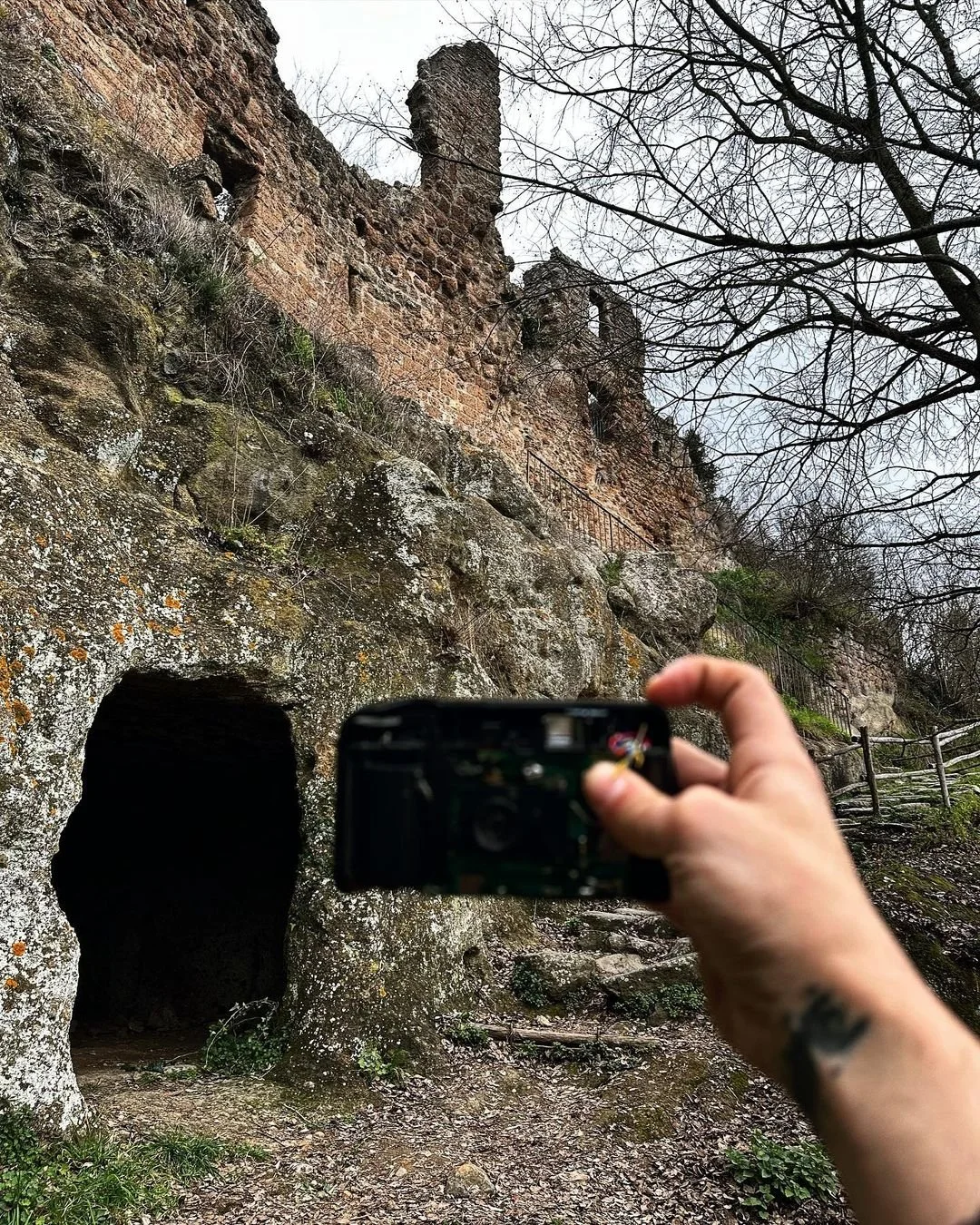 Person taking a photo of a rock cave entrance and an ancient stone structure on a hill, with leafless trees in the background.