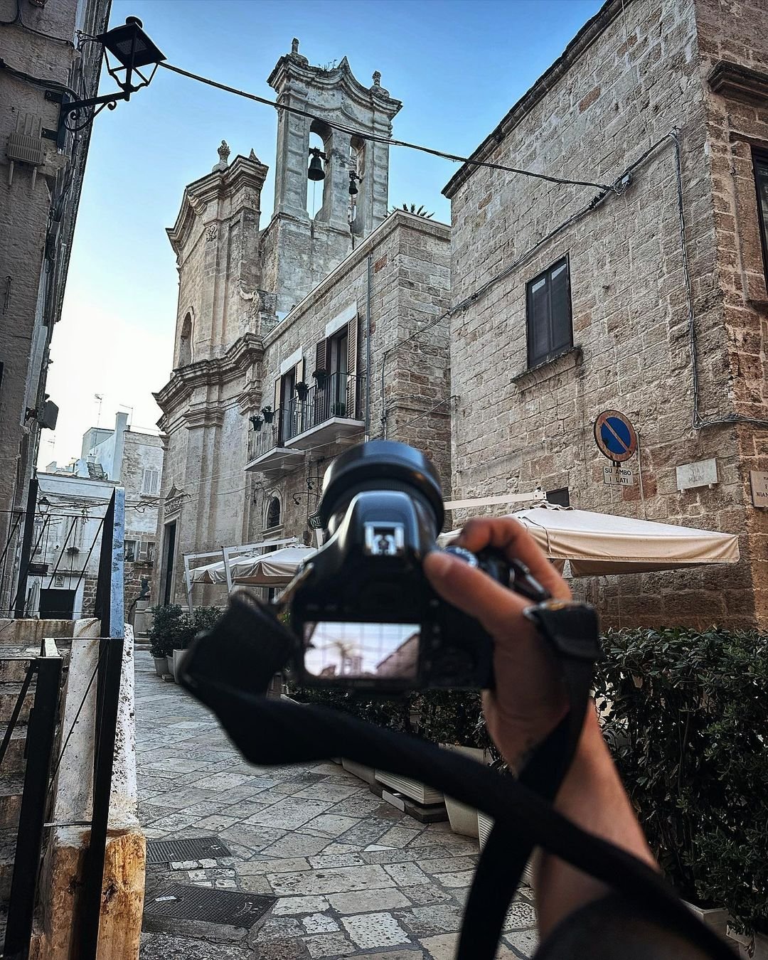 Person taking a photo of an old stone building with a bell tower, narrow cobblestone street, and outdoor umbrellas in a historic European city.