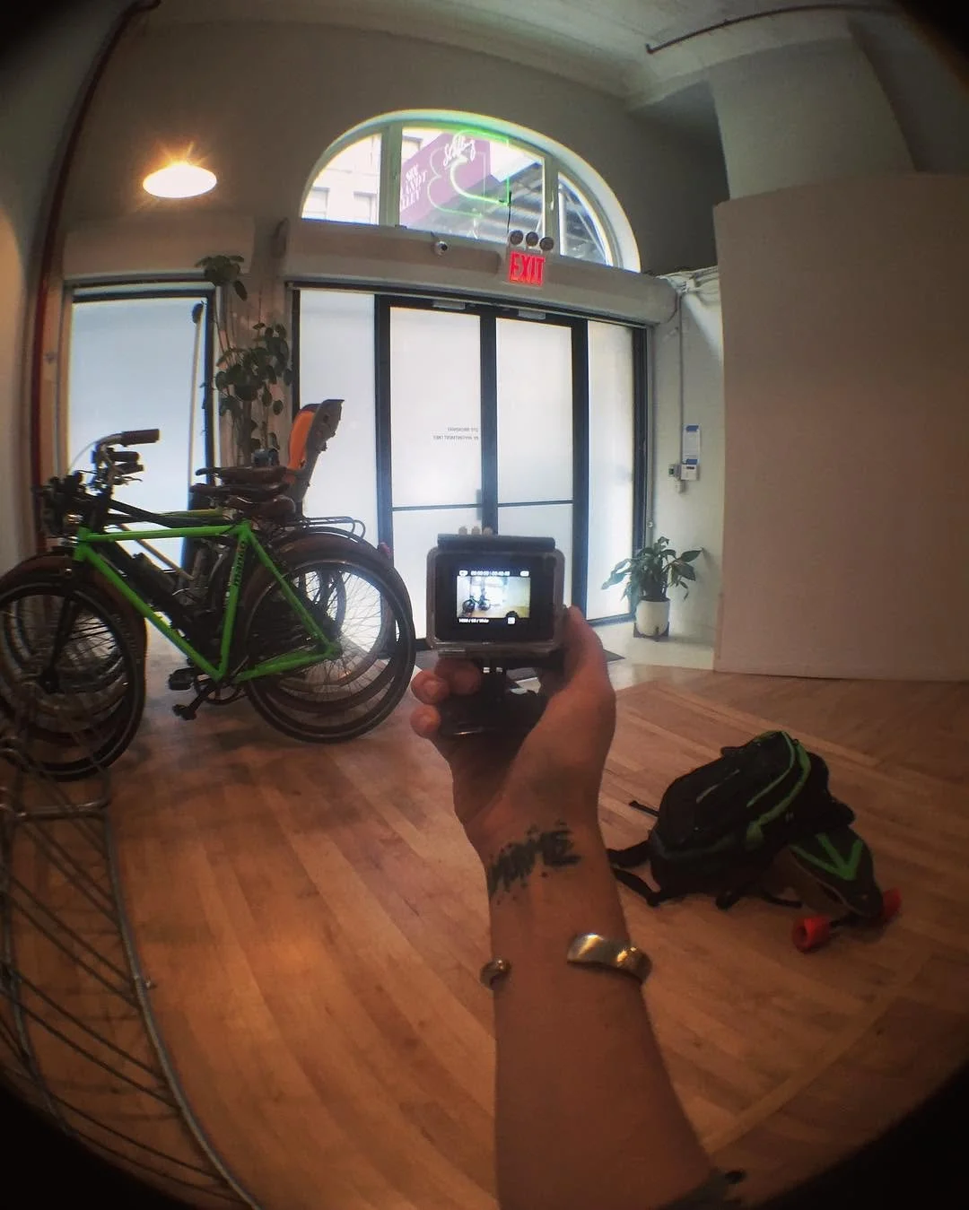 Interior of a bike shop or fitness studio with a green bicycle, a black skateboard with green accents, and a black and green backpack on a wooden floor. A person is holding a camera, taking a photo of the scene.