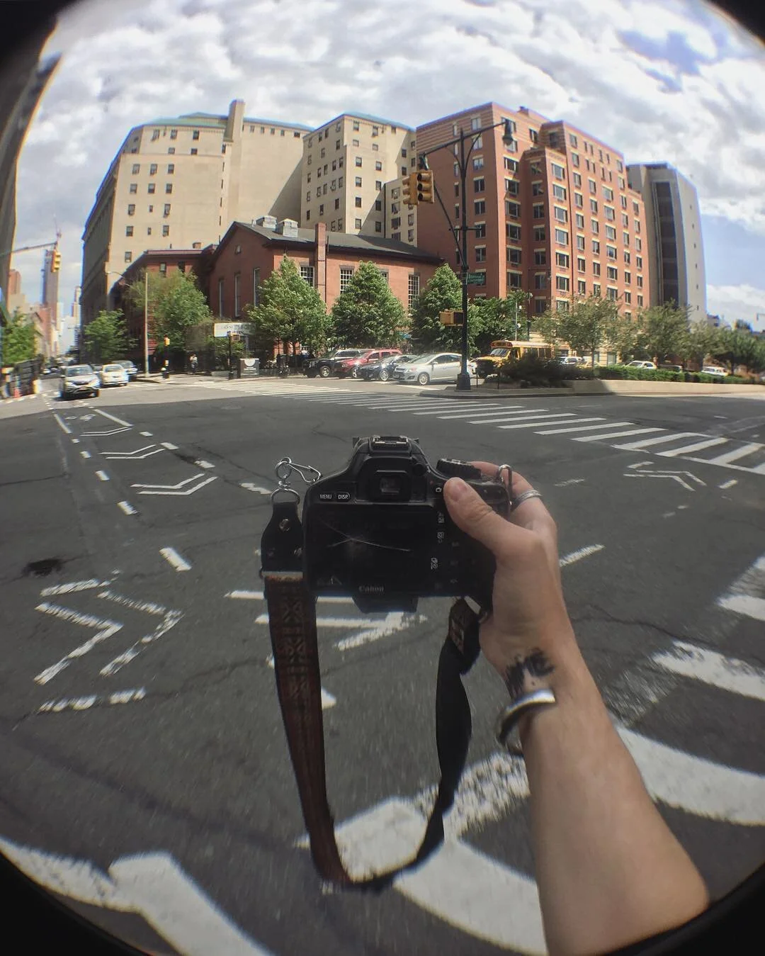Photo taken through a fisheye lens showing a person holding a camera, capturing a cityscape with multi-story buildings, trees, parked cars, and a traffic light at an urban intersection on a partly cloudy day.
