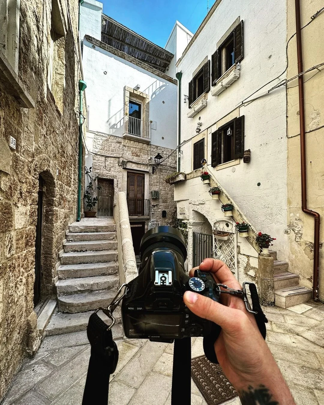 A person holding a camera in a narrow, stone-paved alley with stone and white stucco buildings on either side, featuring small stairs and windows with shutters, potted plants, and decorative elements.