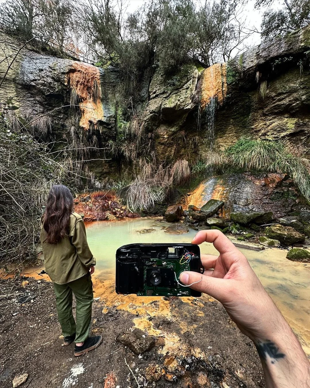 A person in the foreground holding a camera aimed at a woman standing near a small waterfall on a rocky, moss-covered riverbank in a natural outdoor setting.
