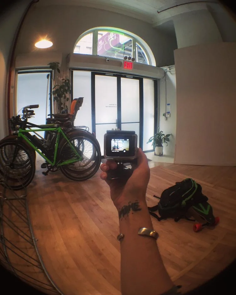 Person holding a camera taking a photo of the room interior, which includes a green bicycle, a backpack, skateboard, plants, large frosted glass door, and neon signs outside.