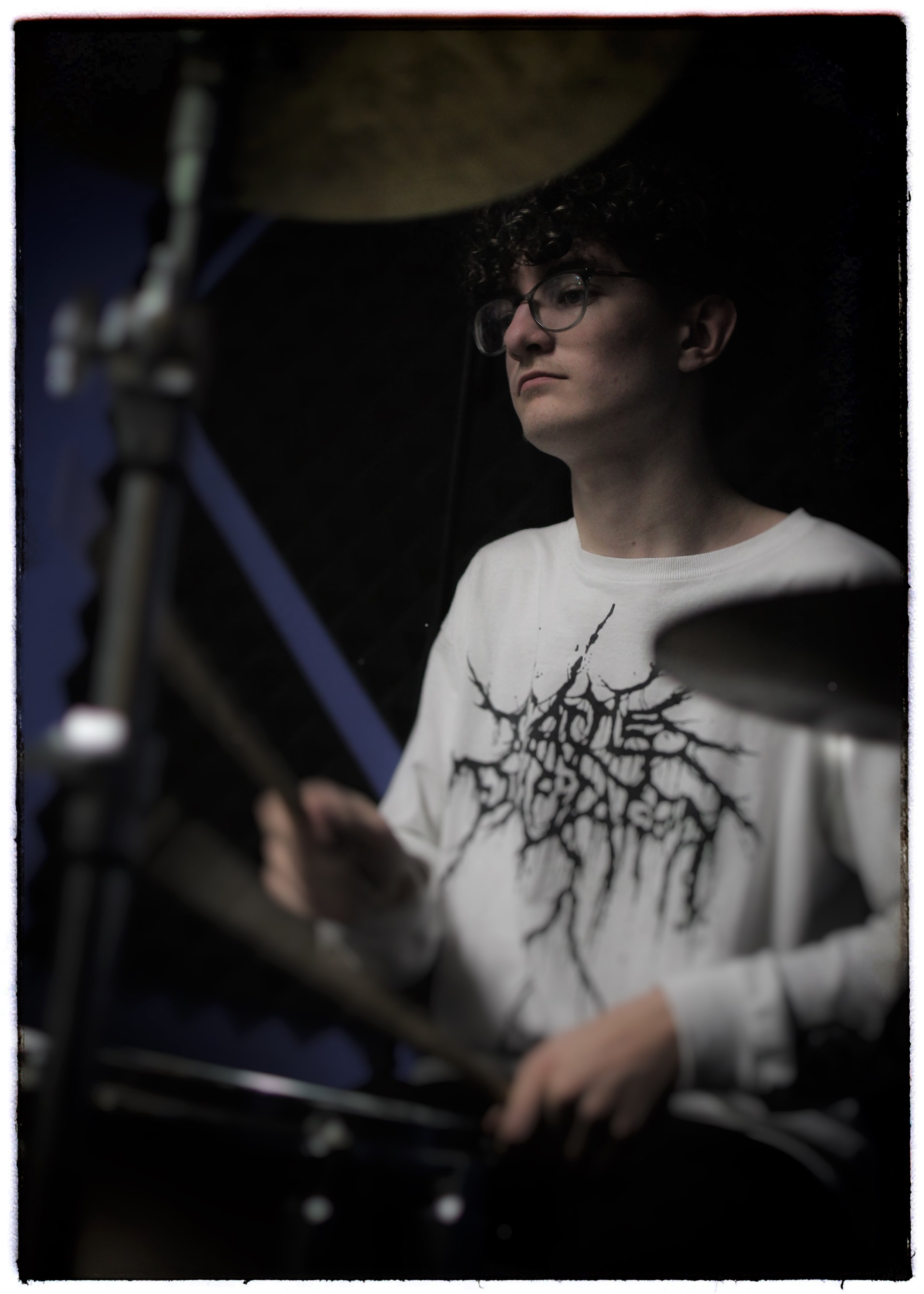 A person with curly hair and glasses playing a guitar or bass guitar, wearing a white shirt with black abstract tree design on the front.