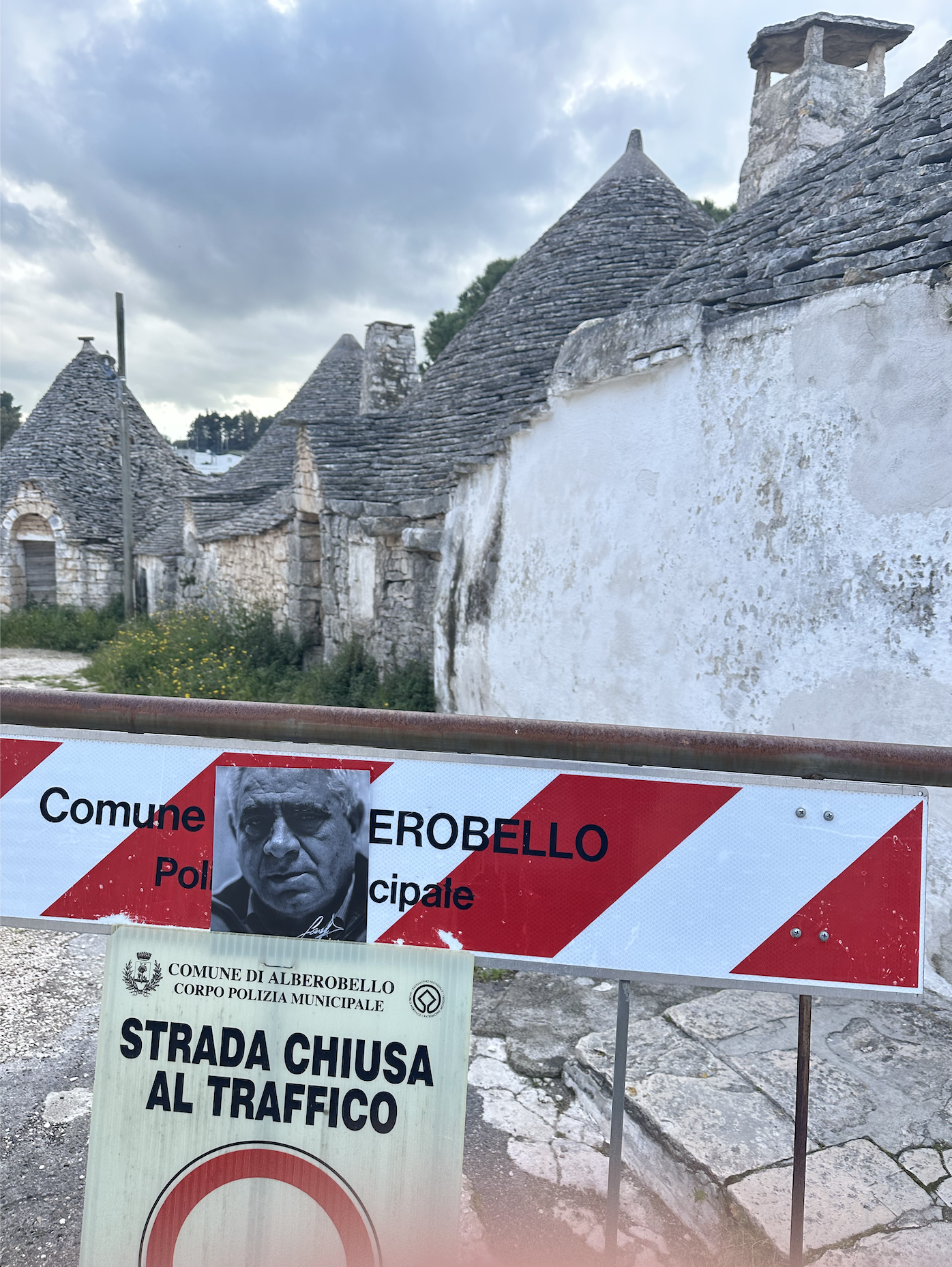 Several warning signs and notices are posted in front of traditional stone buildings with conical roofs, indicating a closed road to traffic, a political campaign poster, and local police information in Italian, with cloudy sky above.