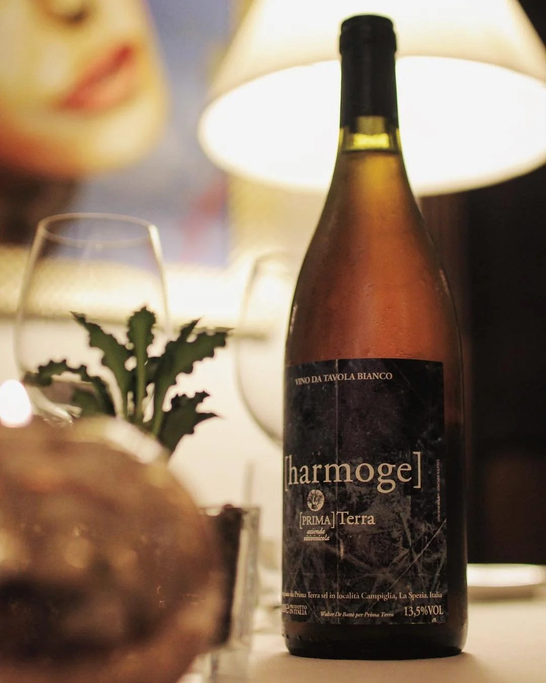 A bottle of Charmoge wine, partially filled, on a table with wine glasses, a lamp, and a small potted plant with spiky leaves, in a cozy, warmly lit setting.
