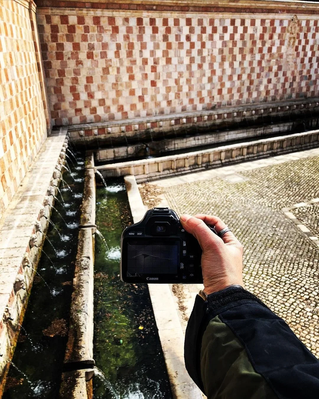 A person holding a camera in front of a water fountain with brick walls and cobblestone pavement.