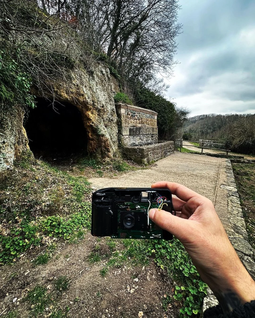 A person holding an open camera pointing towards a rocky cave entrance and a stone wall along a pathway in a natural outdoor setting with trees and cloudy sky.