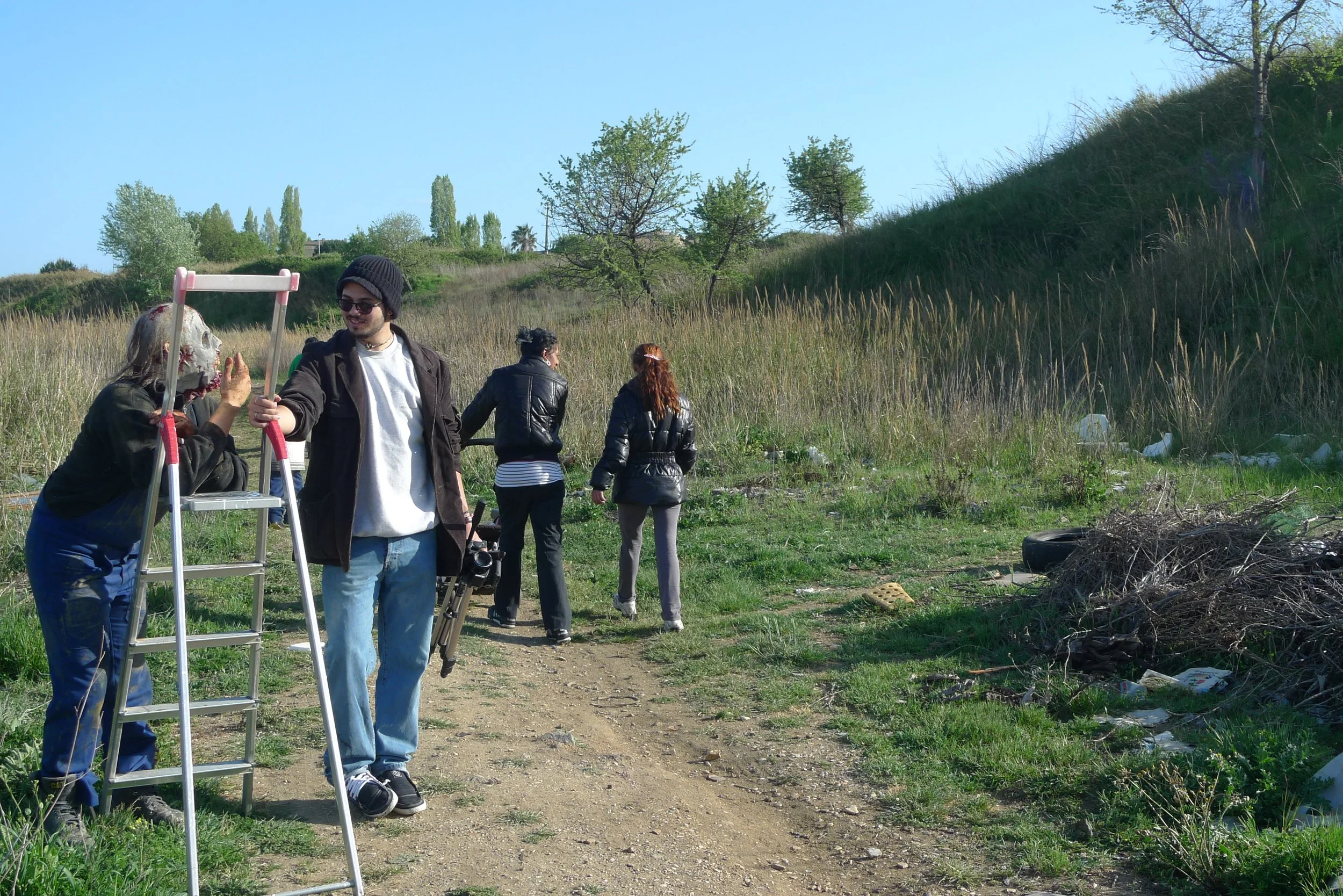 Group of people standing outdoors in a grassy area with trees on a sunny day, one person dressed as a skeleton with a person holding a ladder.