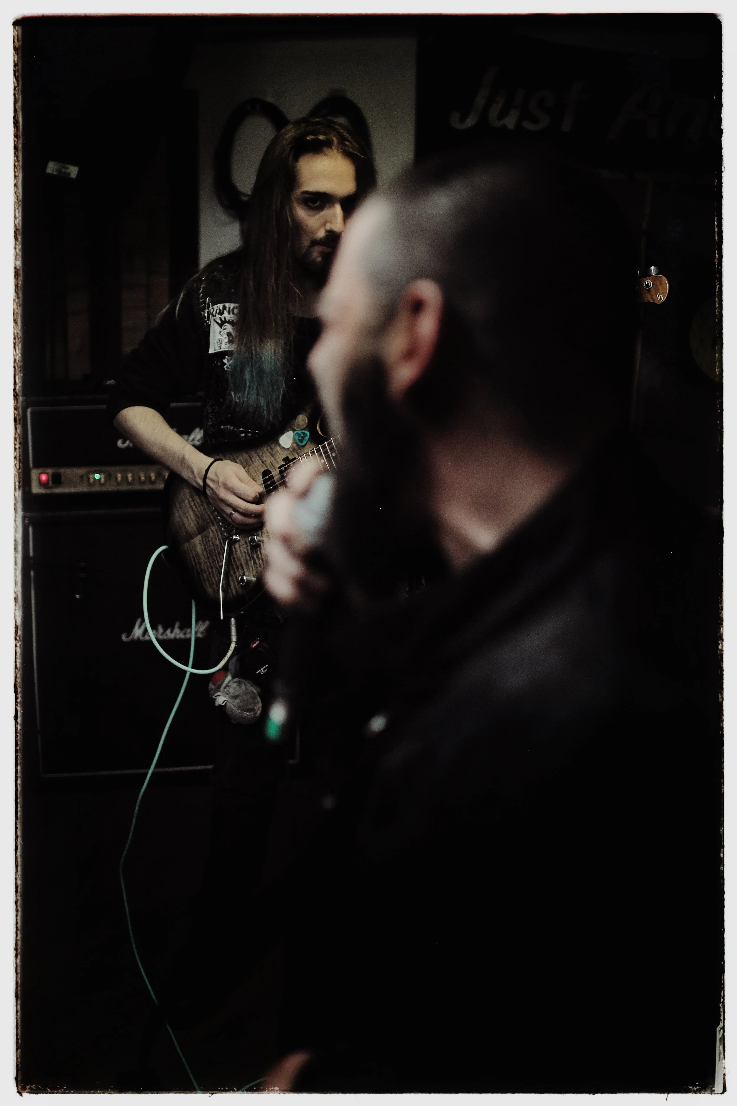 A man with long hair holding a guitar, with a focused expression, in a dimly lit room. Blurry person in the foreground.