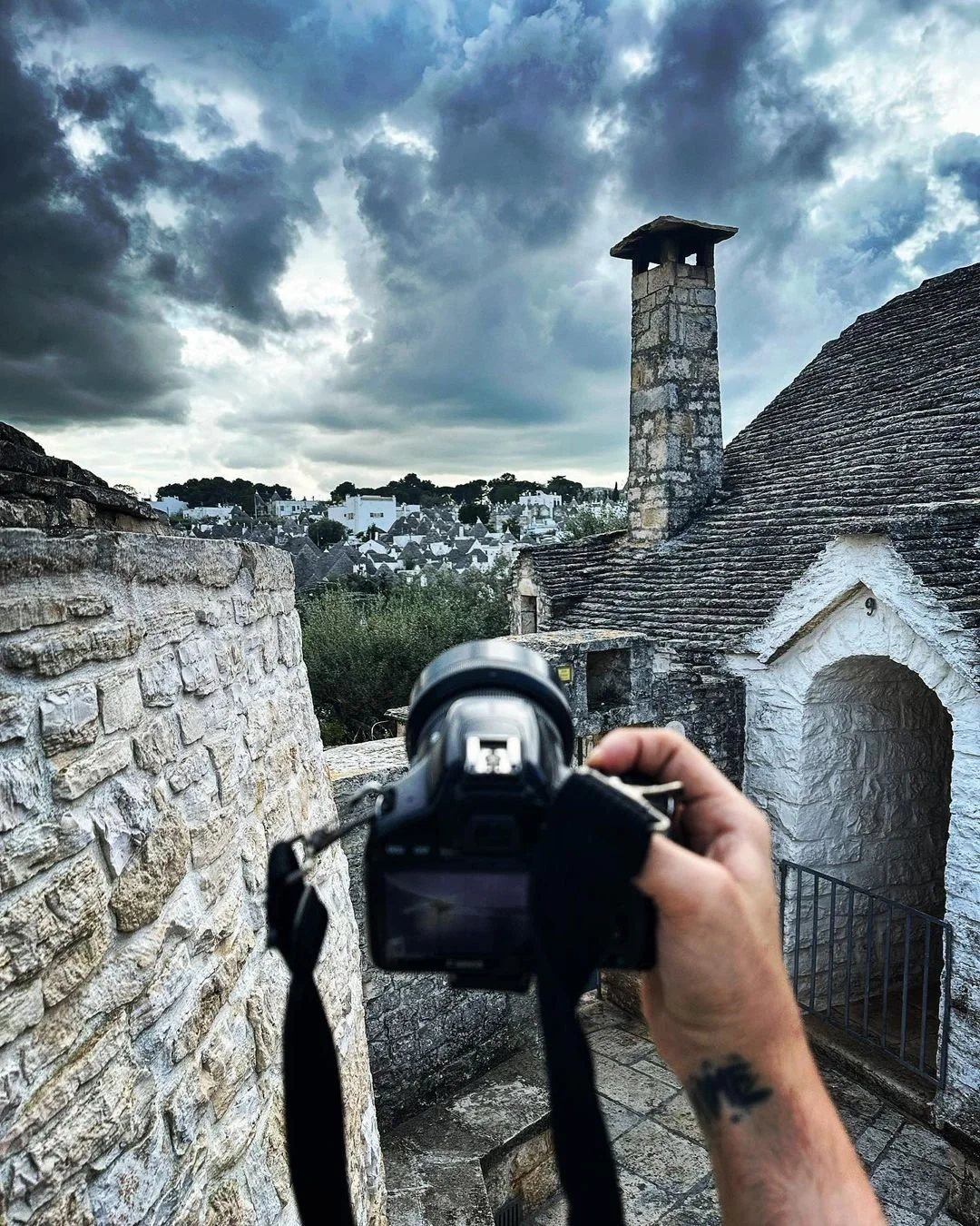 A person holding a camera takes a photograph of a stone building with a chimney, overlooking a town with white houses and a cloudy sky in the background.