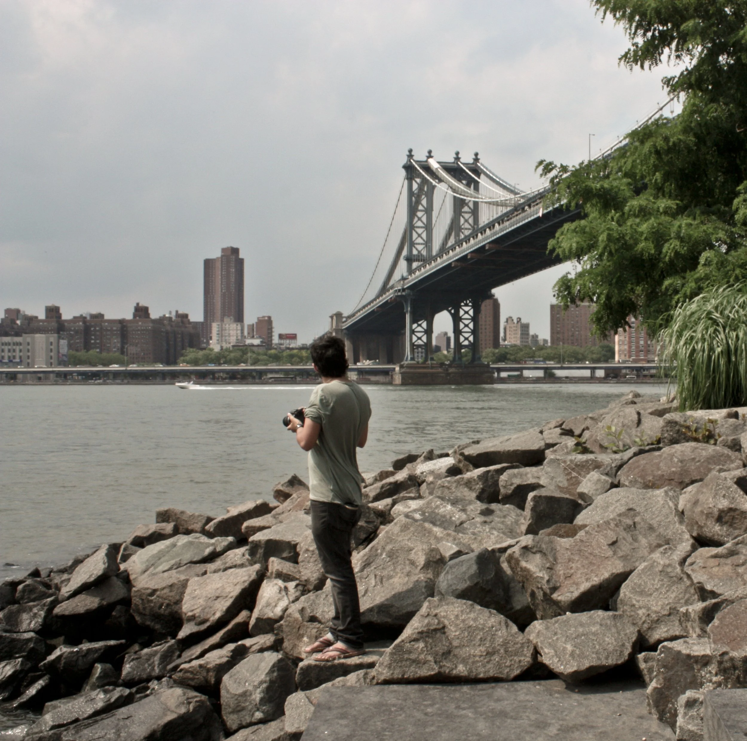 Person standing on rocks by the water with the Manhattan Bridge and city skyline in the background.