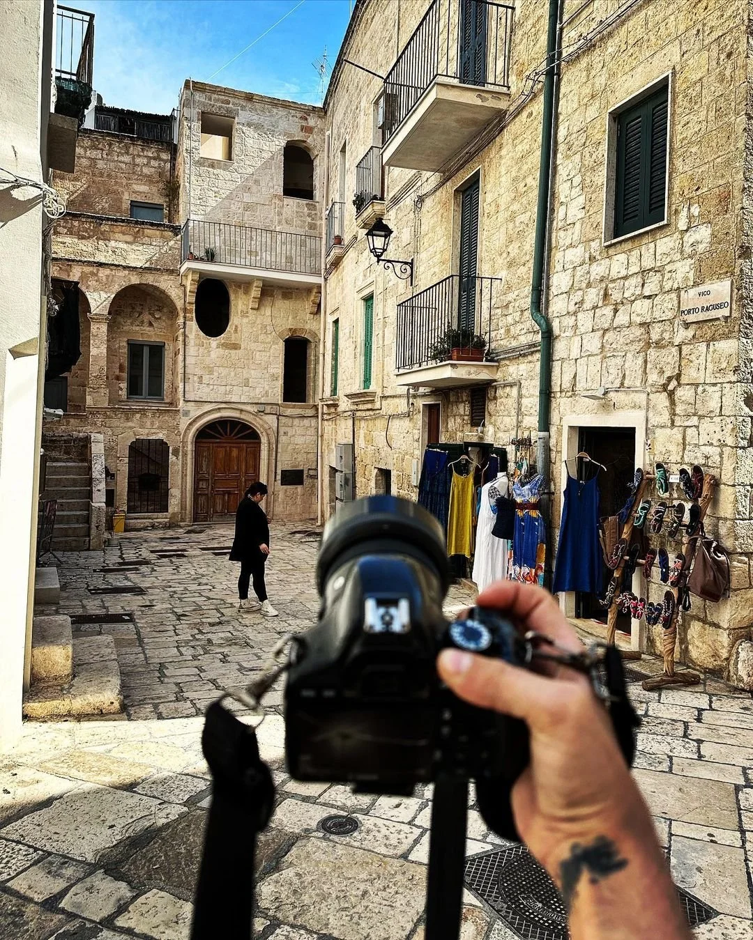 Photo of a photographer taking a picture of a person walking in a historic stone-paved street with old buildings, balconies, and street-side vendors.