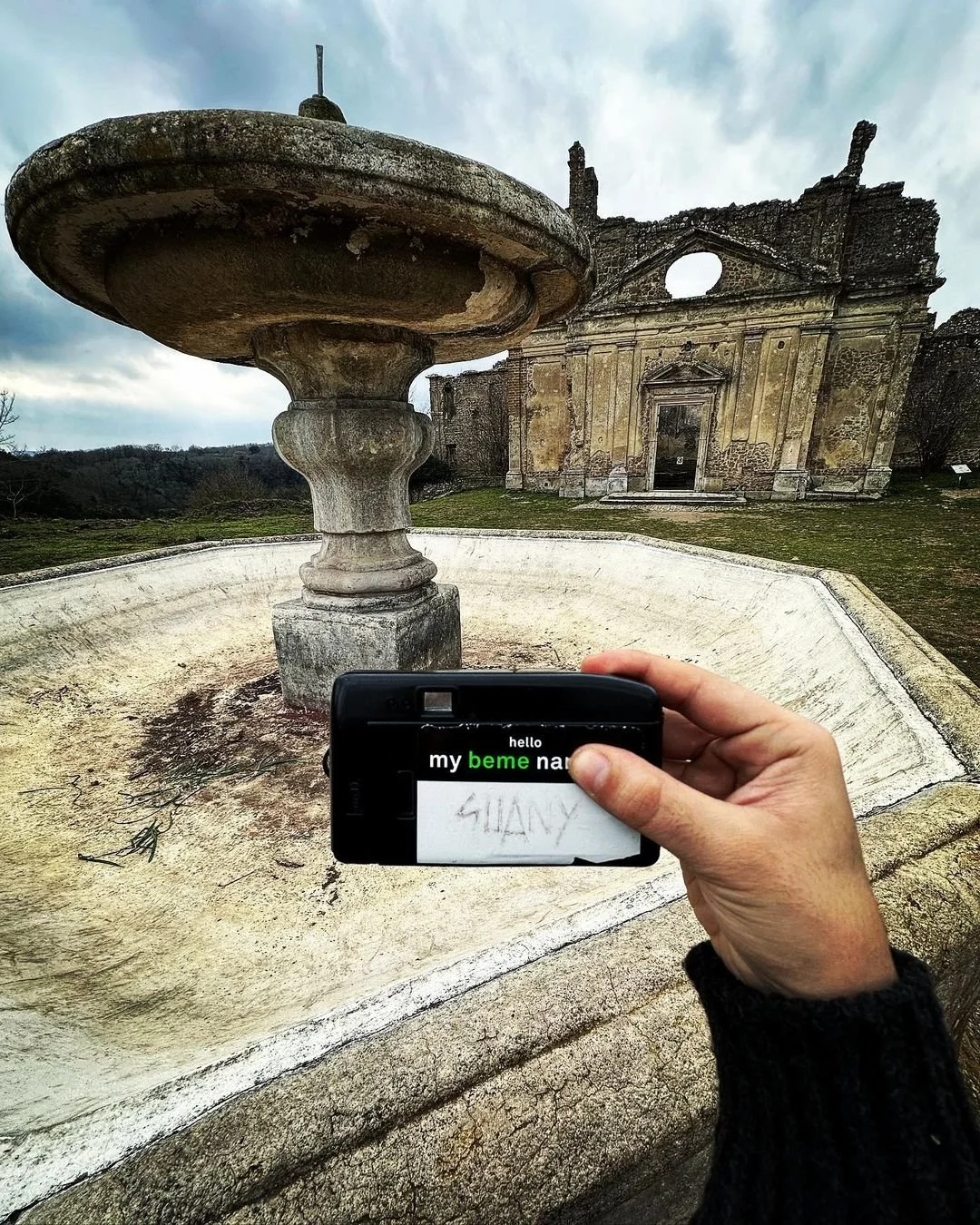 A person taking a photo of an old, weathered stone fountain with a large basin and a decorative top, in front of a ruined historic building with a prominent arch and crumbling walls, under a cloudy sky.