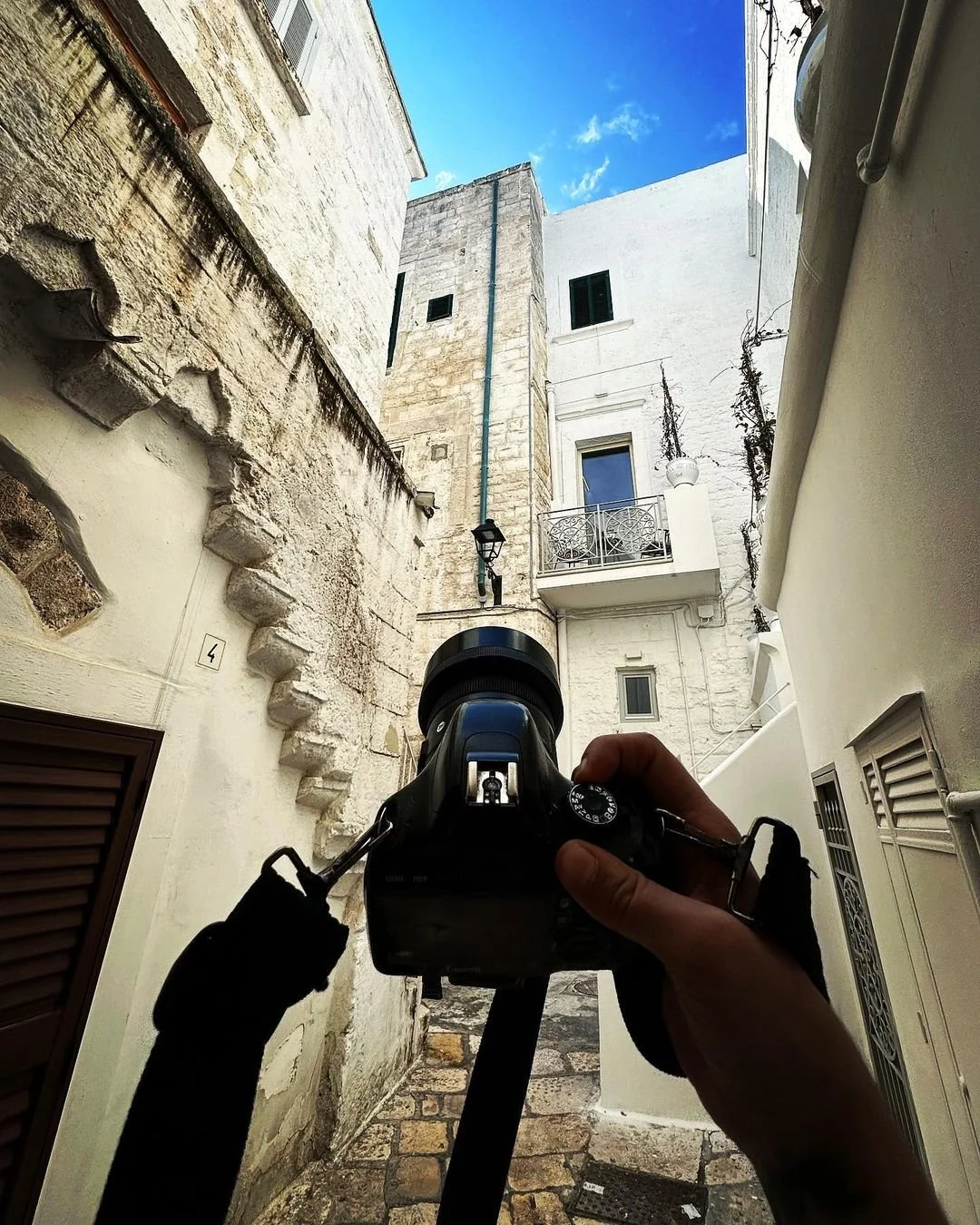 A person holding a camera, taking a photo of a narrow alleyway surrounded by old buildings with white and beige stone walls, some with balconies, against a bright blue sky.