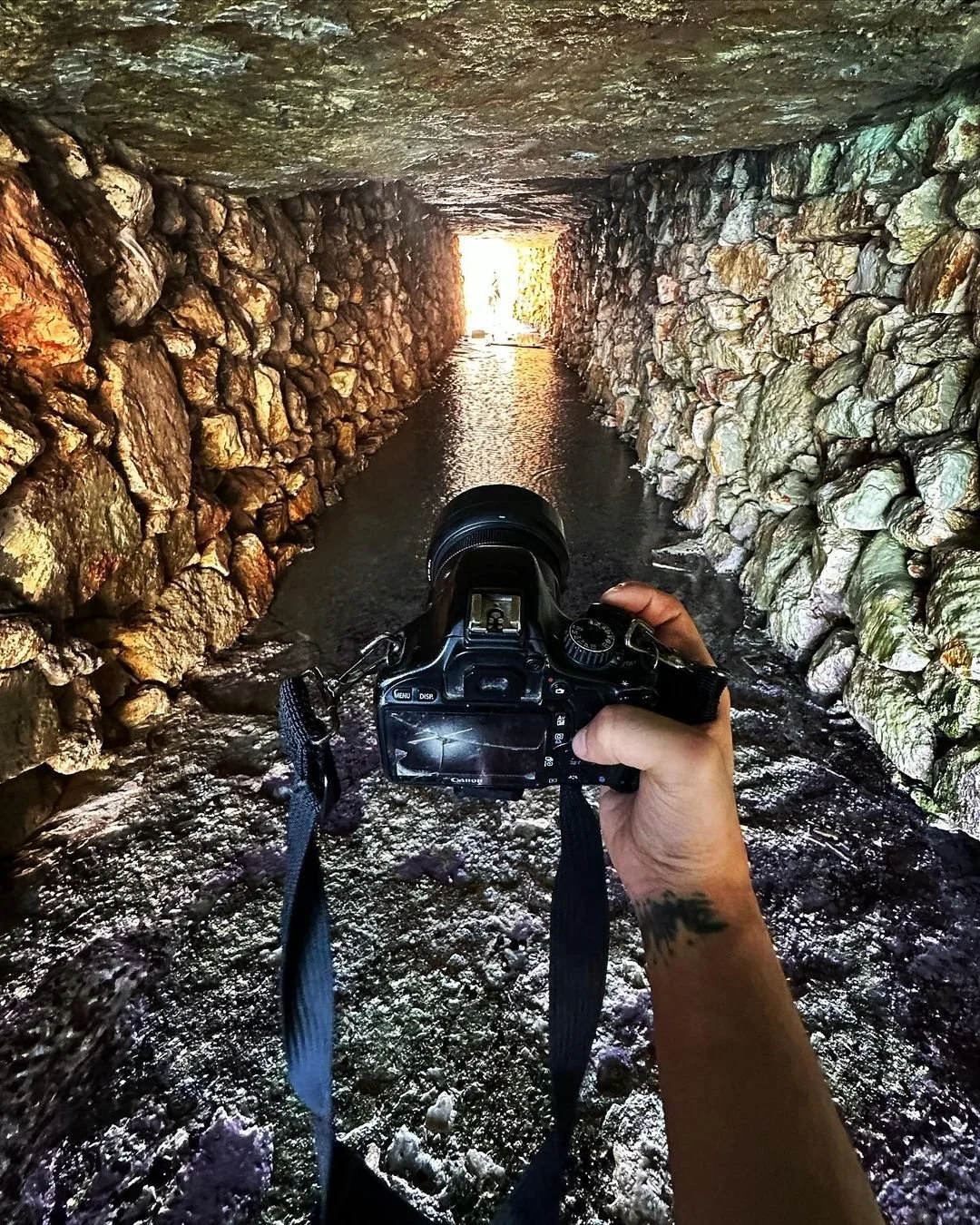 A person holding a camera inside a stone tunnel with water on the ground, leading toward a bright opening at the end of the tunnel.