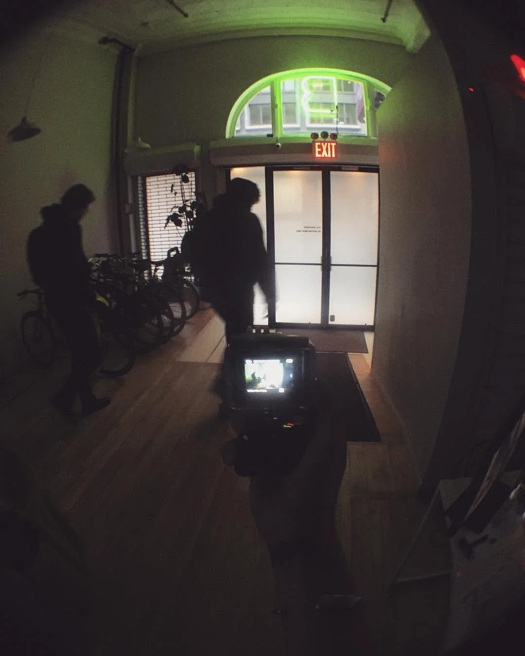 Silhouettes of two people standing inside a dimly lit room near a bicycle rack with several bicycles, glass double doors with an exit sign above, and a neon light in the shape of a dollar sign above the doors.