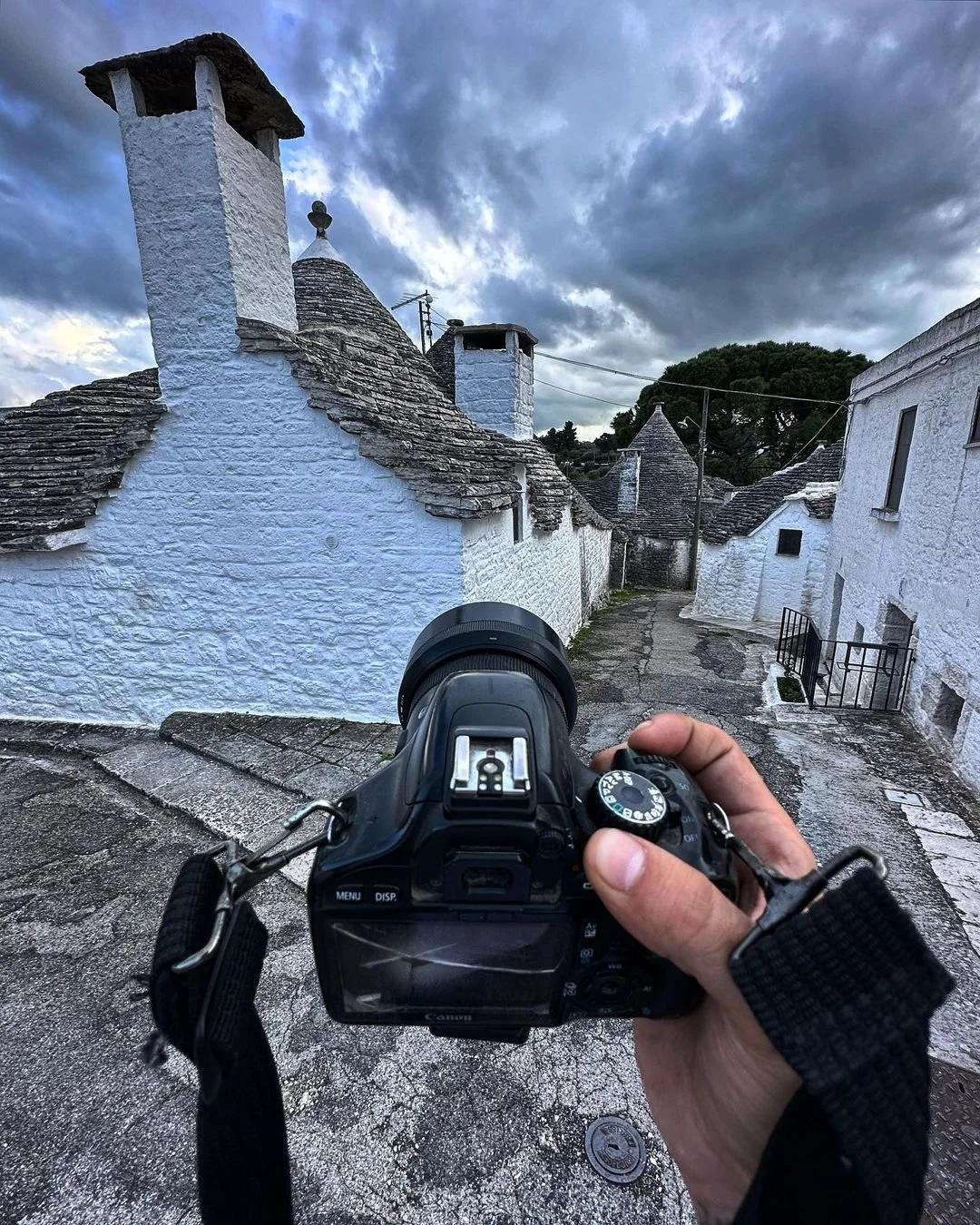 A person holding a black DSLR camera takes a picture of a small village street with white stone houses and conical roofs, under a cloudy sky.